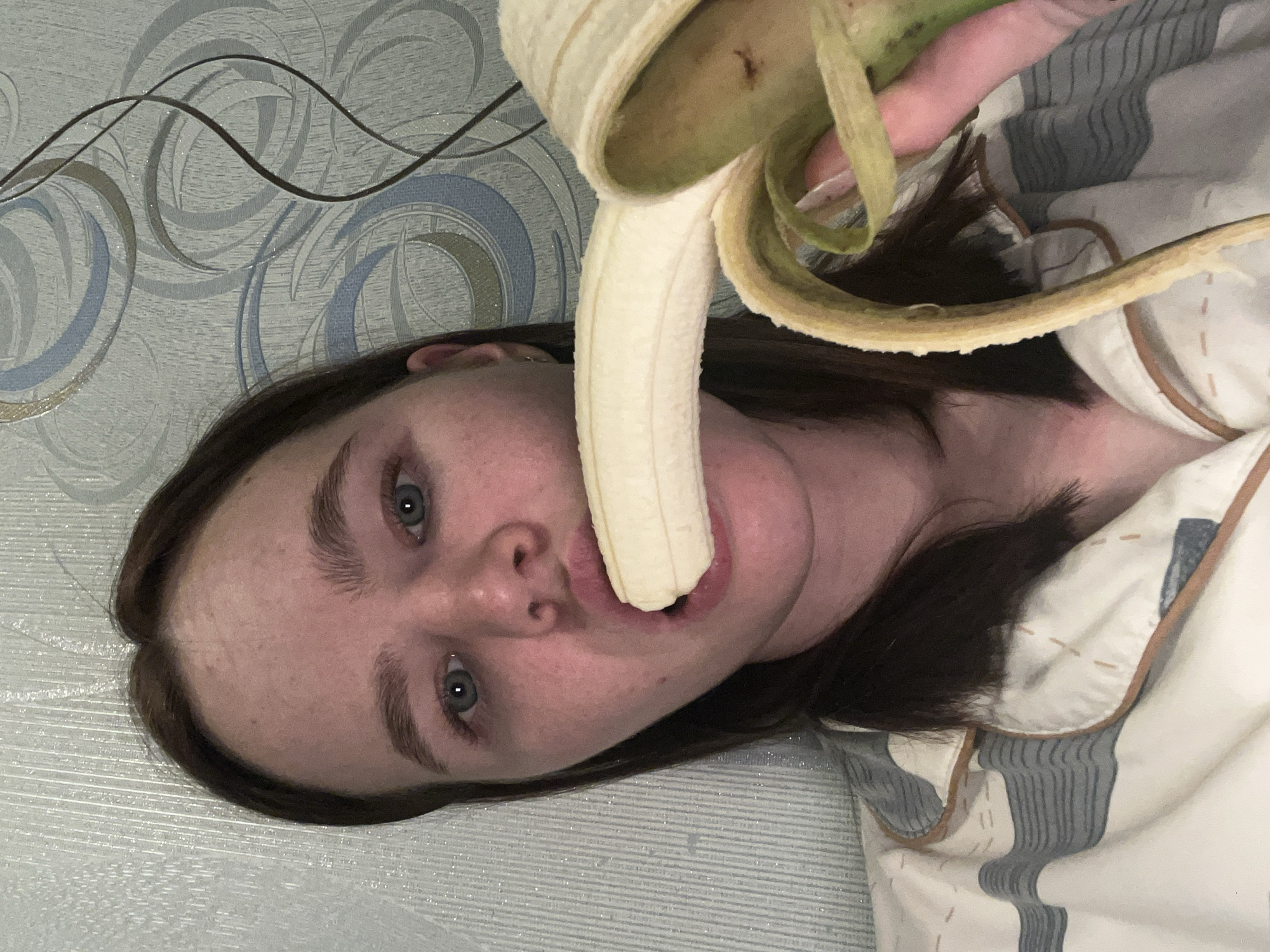 The image, taken in Chaikovsky, Russia, shows a person, appearing to be a young woman, lying on their side in an indoor setting, likely a bedroom. The photograph is oriented sideways. The subject has fair skin, dark brown hair, and blue-green eyes, looking directly at the camera with a neutral expression. A partially peeled yellow banana is held by a hand (likely the subject's) and positioned with its peeled end inside her open mouth.
The subject is wearing what appears to be light-colored pajamas or a top with grey and white stripes. Behind her head and shoulder is patterned wallpaper featuring swirling abstract designs in tones of blue, grey, and hints of gold or brown, interspersed with fine wavy lines. Below her, a patterned bedsheet or blanket with grey and white stripes is visible. No text is visible in the image.