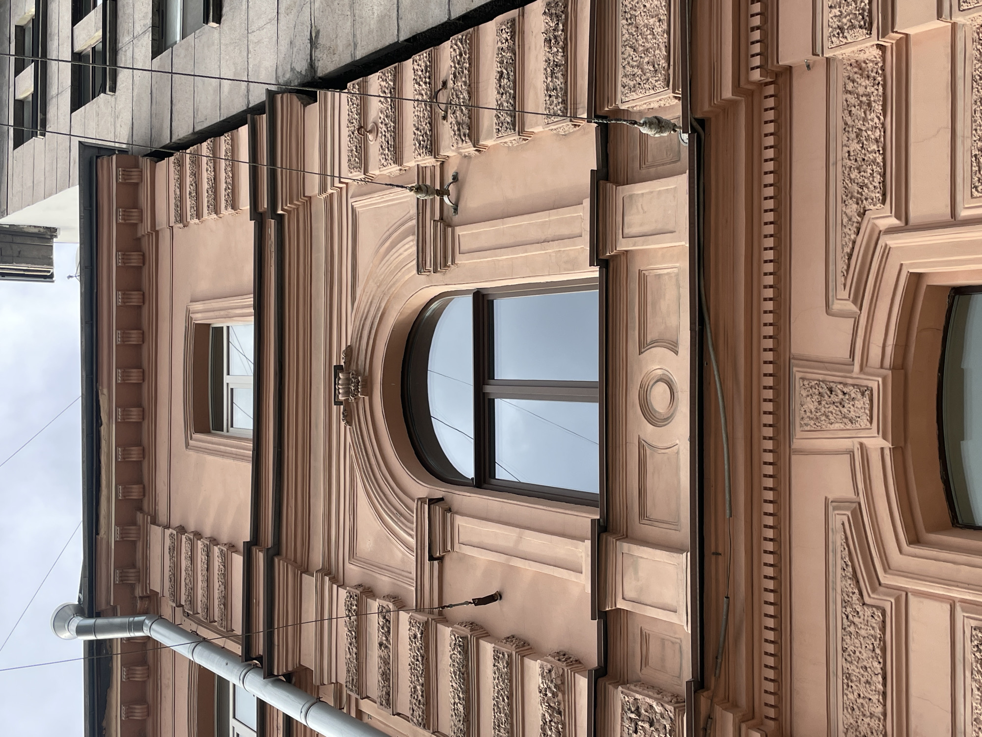 The image shows a fragment of a building facade in Saint Petersburg, Russia. The building is characterized by elaborate architectural details, including framed windows, decorative moldings, and textured panels. The color palette is predominantly light pink with some gray elements. A gutter pipe runs vertically down the left side of the facade. Electrical wiring is strung along the facade.