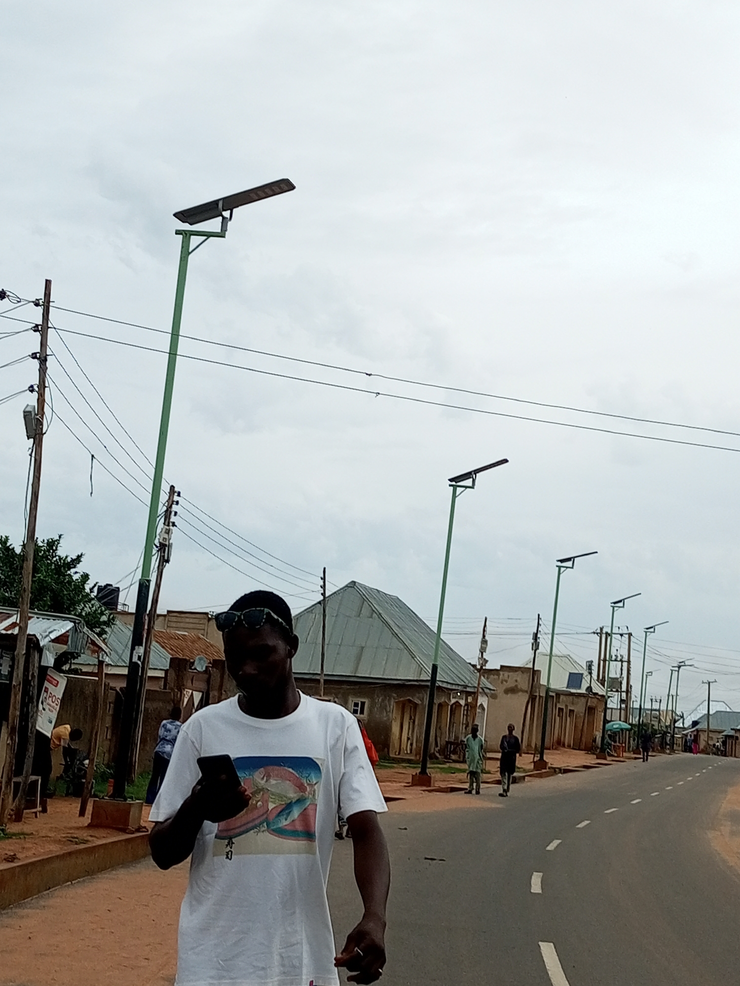 A low-angle, medium shot captures a man wearing sunglasses and a white t-shirt with a graphic print, holding a smartphone in his left hand and walking along a dusty, unpaved road in what appears to be Tempure, Nigeria. The man is the primary subject, positioned in the foreground and slightly off-center to the left. He is looking down at his phone. Behind him, a line of streetlights with solar panels stretches along the road. Several buildings with corrugated metal roofs are visible on the left side of the street. Other individuals are scattered throughout the scene, including two men walking in the middle ground and a few figures further in the distance. The sky is overcast, creating a diffused light. The environment suggests a semi-urban or rural setting with modest infrastructure.