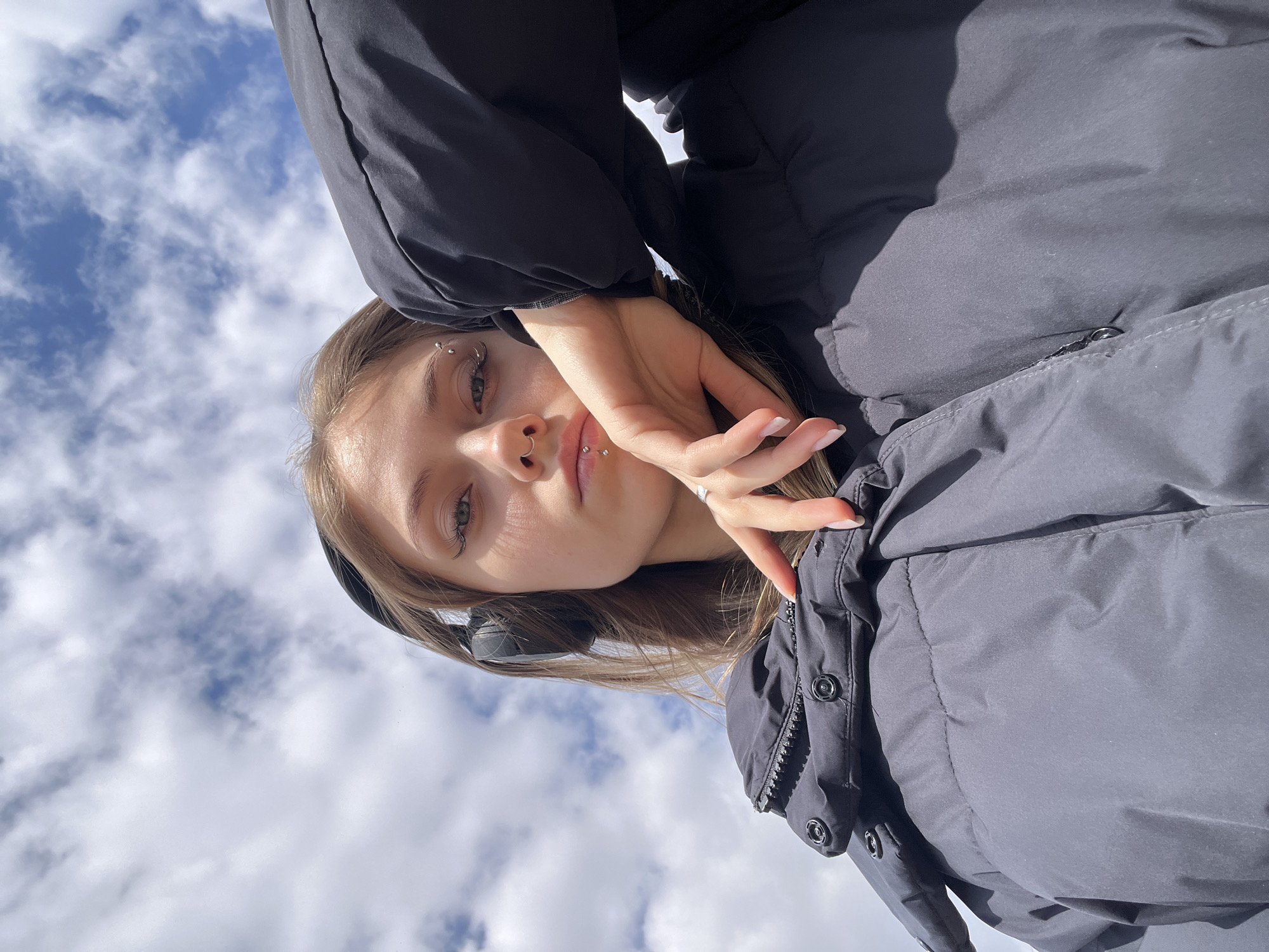 A young woman is depicted in an inverted, low-angle shot against a bright blue sky filled with white, fluffy clouds. She is positioned upside down in the frame, looking directly towards the viewer. Her light brown hair falls around her shoulders, and she is wearing a dark, possibly black, puffer jacket.
Notable features of the woman include several facial piercings: a septum ring, a philtrum piercing, and multiple small barbell piercings above her left eye. She has light-colored eyes and appears to be wearing black over-ear headphones. Her right hand is raised, partially obscuring her face, with her fingers spread slightly and a ring visible on one finger. The lighting suggests a clear, sunny or partly cloudy day outdoors. The overall mood is casual, characteristic of a stylized selfie. No text is visible in the image. This scene takes place in or around Фоки, Russia.