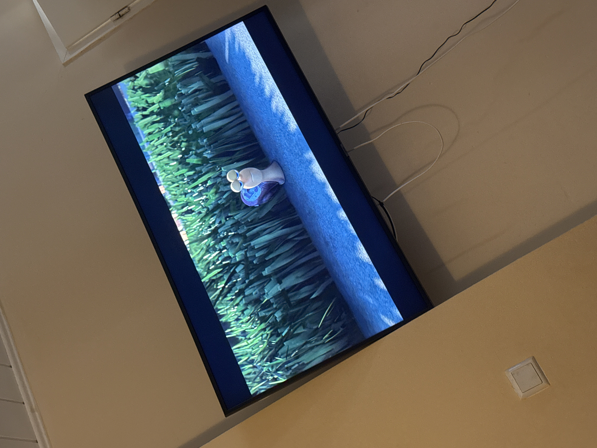 The image shows a television screen displaying a scene from the animated movie "Turbo." The scene features a cartoon snail with large eyes and a blue shell, positioned on a blue surface that appears to be water or a reflection. Behind the snail is a dense field of tall, green grass, with some white highlights suggesting light reflecting off the blades. The television is mounted on a wall, angled slightly downwards. The wall appears to be a neutral, light color, possibly beige or cream. To the left of the television, a square, white light switch is visible on the wall. Several white and black cables are connected to the television and trail down the wall. The overall environment suggests an indoor setting, likely a living room. The location is identified as Ahlen, Germany.