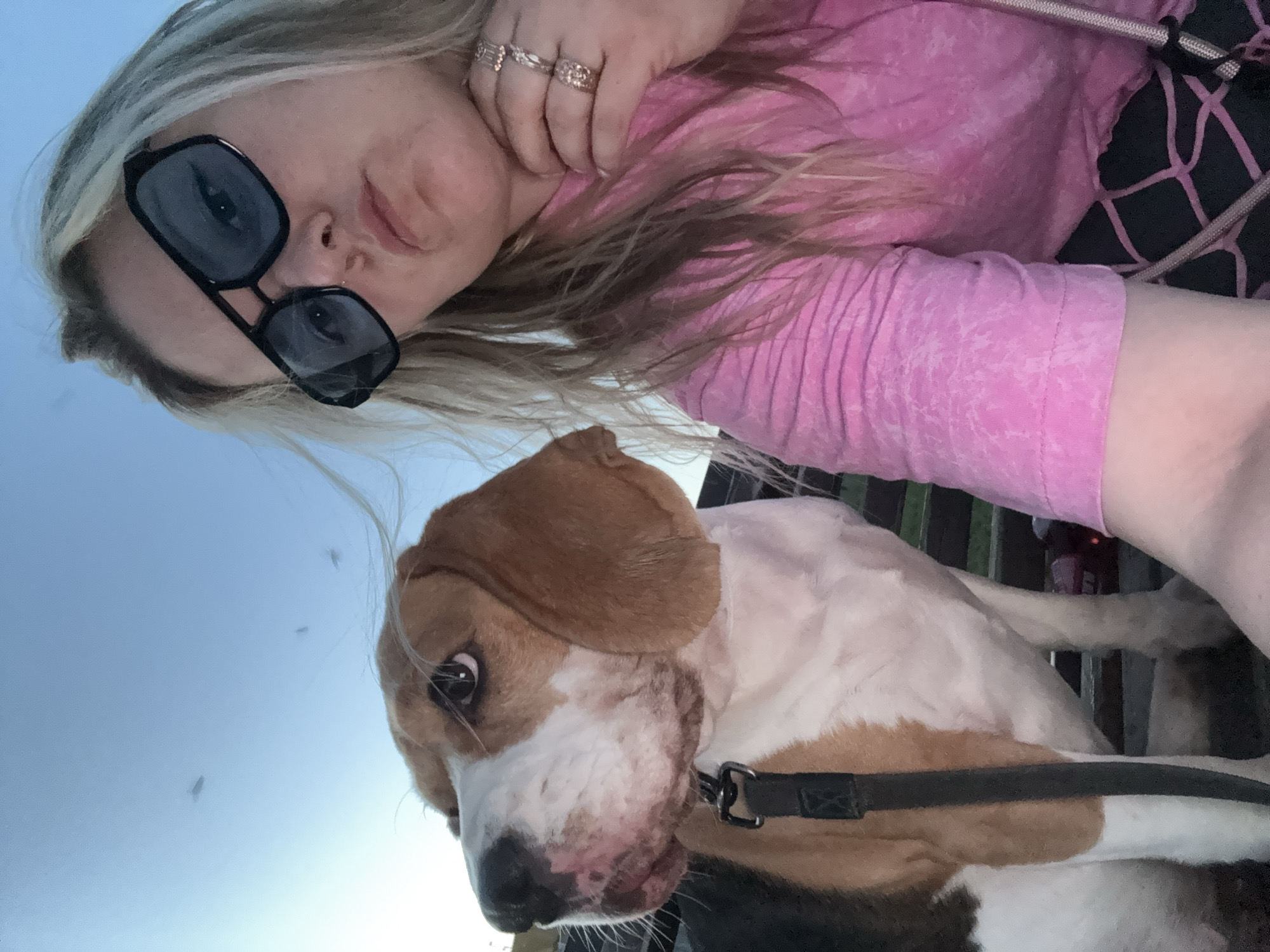 A selfie shows a woman and a beagle. The woman has blonde hair and is wearing sunglasses and a pink shirt. The dog is mostly white with brown patches and is wearing a black collar. The background is blurry and includes a blue sky and some green vegetation. The picture appears to have been taken outdoors. There is no evidence to suggest this picture was taken in Марковский, Russia.