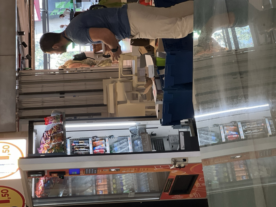This image depicts an indoor scene in San Cristóbal de La Laguna, Spain.
In the left foreground, a white and orange vending machine is present, with its right service door open. The machine's glass front displays rows of bottled beverages (e.g., Fanta, water). The open section reveals internal shelving stocked with bags of crisps/chips on the top shelf and various canned/bottled drinks (e.g., K-Tonic, water) on shelves below. Above the vending machine, an illuminated sign displays "1,50" in red and orange text.
To the right, a male individual is visible from the mid-torso to the lower legs, facing left. He has short, dark hair and a beard/stubble. He wears a blue short-sleeved t-shirt, light beige trousers, and a dark-colored smartwatch on his left wrist. His hands are slightly cupped, positioned as if holding or preparing to handle an item. On the floor in front of him are two dark blue plastic crates containing various cardboard boxes and packing materials. Behind these crates, several white chairs are stacked in a disorganized manner, alongside a light-colored table.
In the background, an accordion-style security grate is partially visible, behind which an open doorway leads to an exterior or brightly lit internal area. A female individual, viewed from behind, wearing light-colored patterned trousers and a dark backpack, stands near this doorway. To her right, a second male individual wearing a white shirt is partially visible in the background.
The lower third of the image consists of a clear, inverted reflection of the vending machine, the blue crates, and the lower half of the primary male subject, indicating a highly reflective floor or surface in the immediate foreground.
