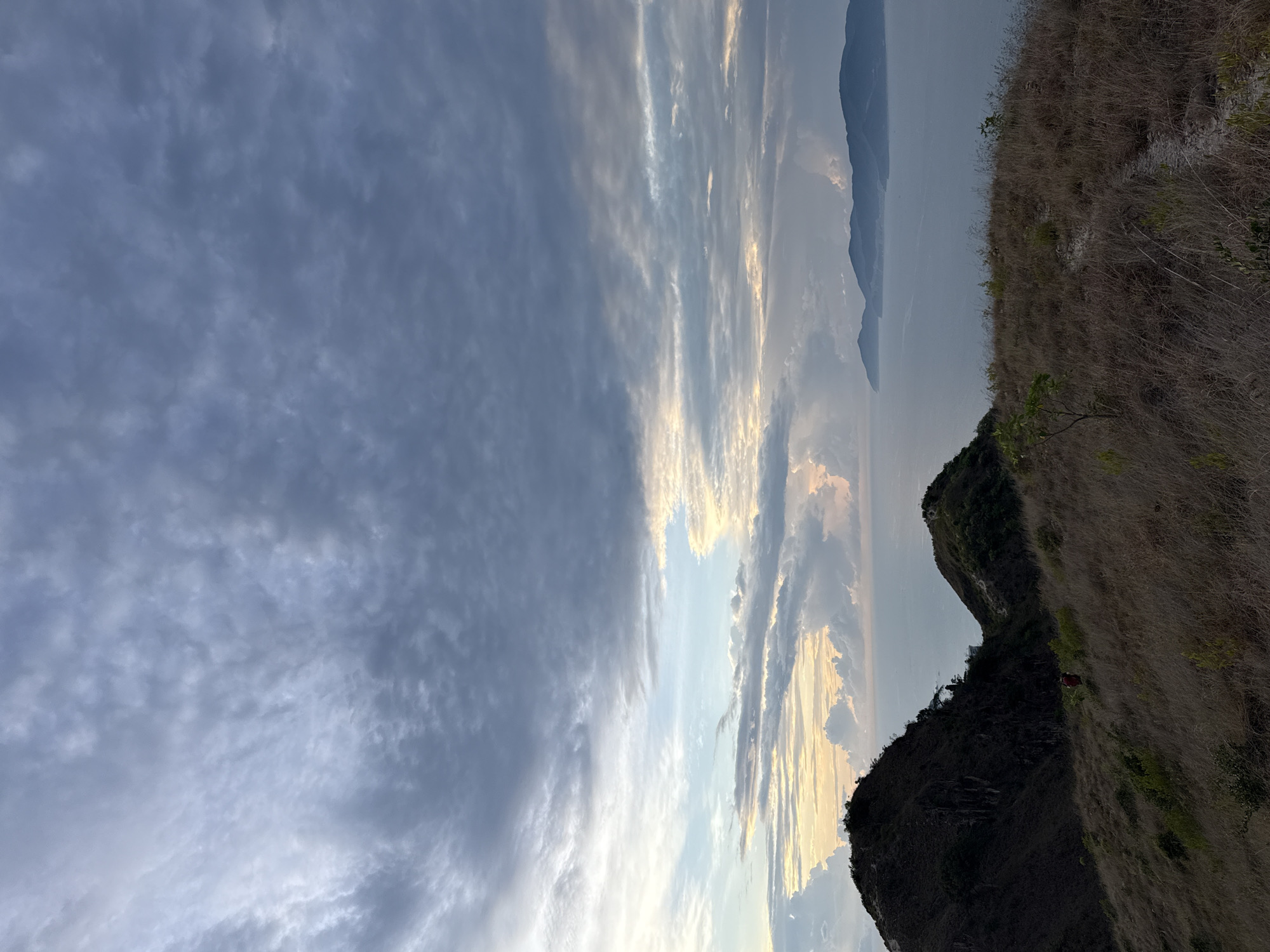 The image shows a landscape in Bac Nha Trang Ward, Vietnam. The right side of the frame is dominated by a steep, grassy hillside, partially covered in dry, brown vegetation. A rocky outcrop or cliff face is visible at the base of the hillside. In the background, beyond the hillside, is a body of water with mountains in the distance. The sky above is filled with clouds, ranging from dark gray to lighter shades, with patches of blue visible. The clouds appear to be illuminated by the setting or rising sun, creating a soft, golden glow in some areas. The overall scene appears to be a landscape captured during dusk or dawn.