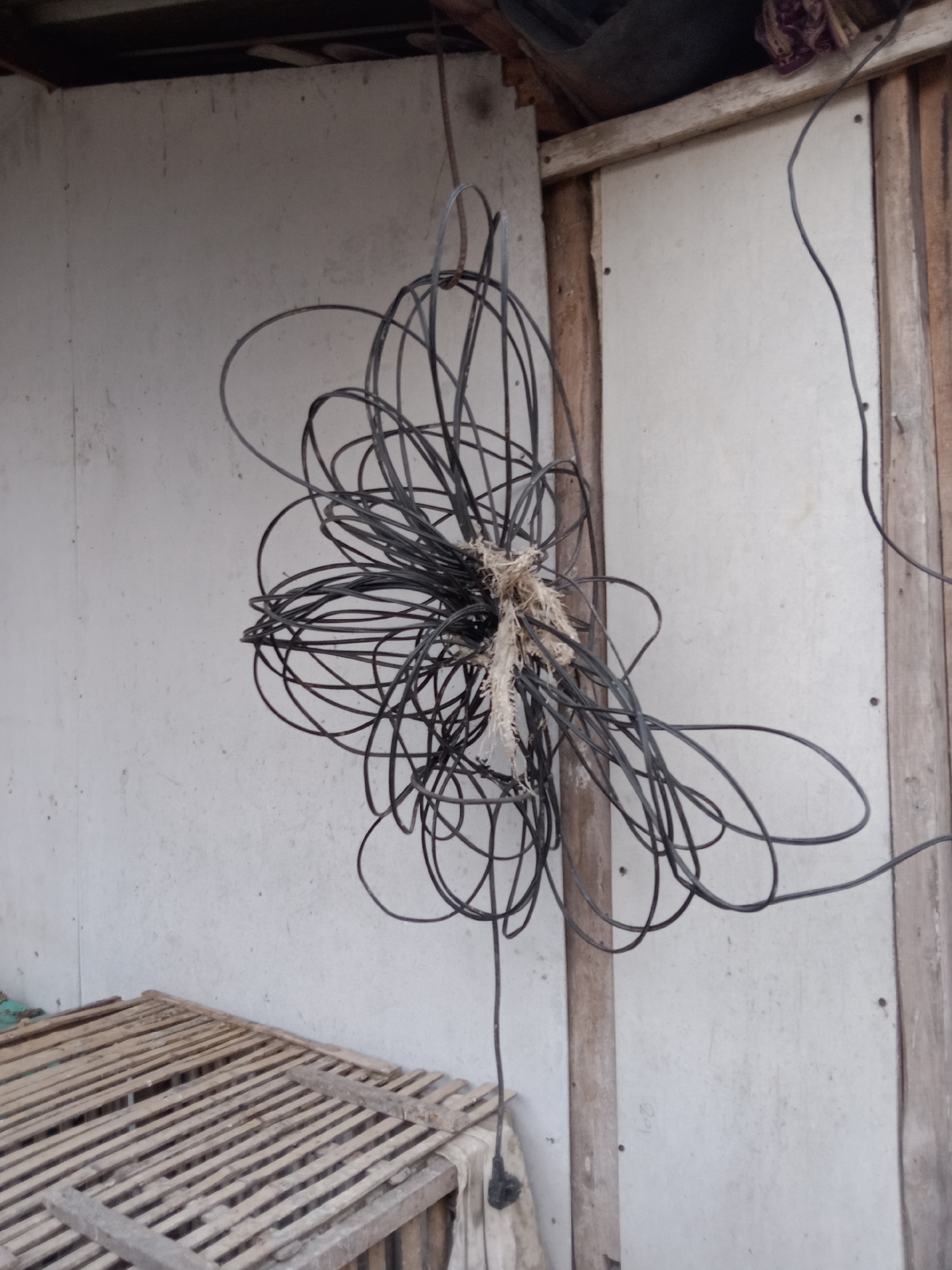 In the city of Jumput, Indonesia, a tangled mess of black electrical wires hangs suspended in front of a lightly textured, off-white wall. The wires, bound together with a rough, fibrous material, form a chaotic knot. To the left of the wires, a rustic wooden structure, possibly a cage or platform made of slats, is visible. The background wall appears to be part of an outdoor structure, with wooden beams and framing visible on the right side. The lighting suggests an overcast day or a shaded area.