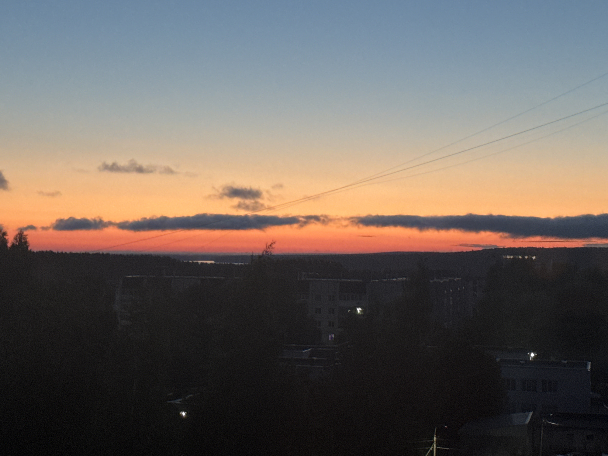 The image depicts a panoramic view of a city during twilight, likely Chaikovsky, Russia, given the context of provided crops. The sky transitions from a pale blue at the top to a vibrant orange and red near the horizon, indicative of a sunset. Faint, wispy clouds are scattered across the sky, and three distinct contrails from aircraft are visible diagonally across the upper right quadrant. Below the sky, a dark, silhouetted landscape stretches across the middle ground. This landscape features a dense line of trees bordering a developed area. Within this developed area, several multi-story buildings, characteristic of Soviet-era apartment blocks, are discernible in silhouette. Some windows in these buildings are illuminated, suggesting internal lighting. The foreground is dominated by dark, shadowy trees, obscuring any ground-level details. The overall atmosphere is serene and nocturnal, with the lingering colors of the sunset providing the primary illumination.