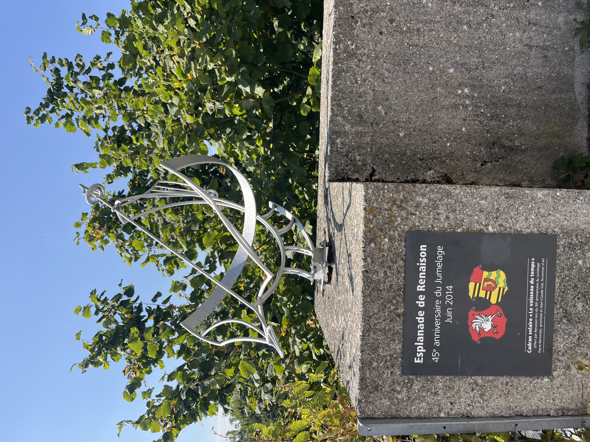 The image shows a metal sundial sculpture placed on top of a concrete block. The sculpture is abstract and silver-colored, with curved metal pieces and a gnomon. A dark plaque is affixed to the front of the concrete block, featuring text and two coats of arms. The text on the plaque reads "Esplanade de Renaisson 45e anniversaire du Jumelage Juin 2014" and "Cadran solaire « Le vaisseau du temps » Offert par Renaîson lors du 30e anniversaire du Jumelage Pierre Remontet, gnomiste et Jean-Claude Vial, ferronnier d'art". This indicates the sundial is named "Le vaisseau du temps" and was offered by Renaîson for the 30th anniversary of the twinning. The background consists of lush green foliage and a clear blue sky. The concrete block appears to be part of a larger structure, possibly a monument or a garden feature. The location is stated to be Epagny, Switzerland. There are no people or animals visible in the image. The scene is well-lit, suggesting a sunny day.