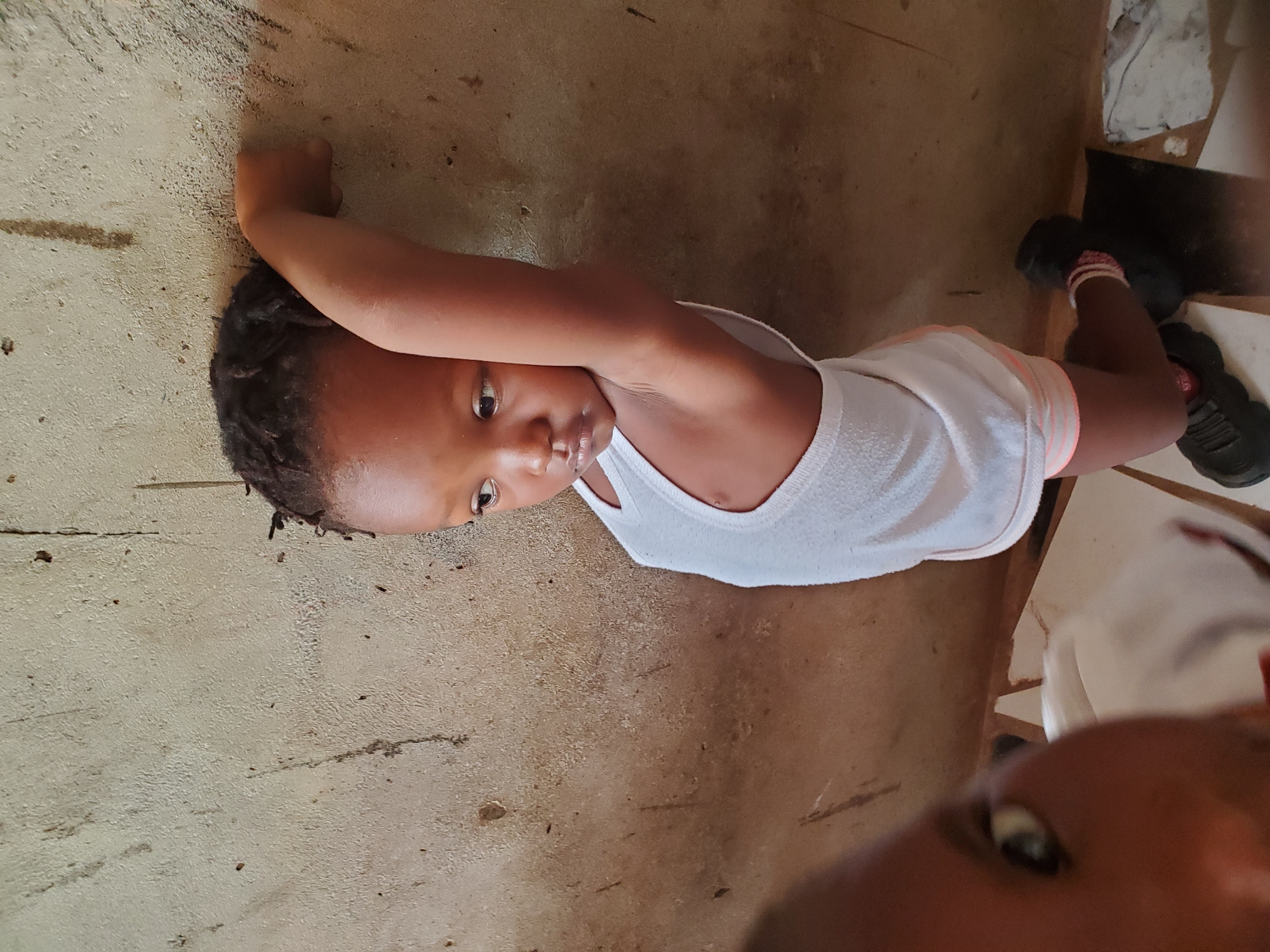 A child, likely a toddler, is lying on their back on a painted concrete or cement floor in Ukwu-Abwa, Nigeria. The child is wearing a white tank top and shorts with a pink and white striped trim. Their right arm is raised and bent at the elbow, with their fist near their head. They are wearing black athletic shoes with pink socks. Another person’s face is partially visible in the lower right corner of the frame. Part of a white object and a dark surface are in the upper right corner, possibly a small shelf or stand.