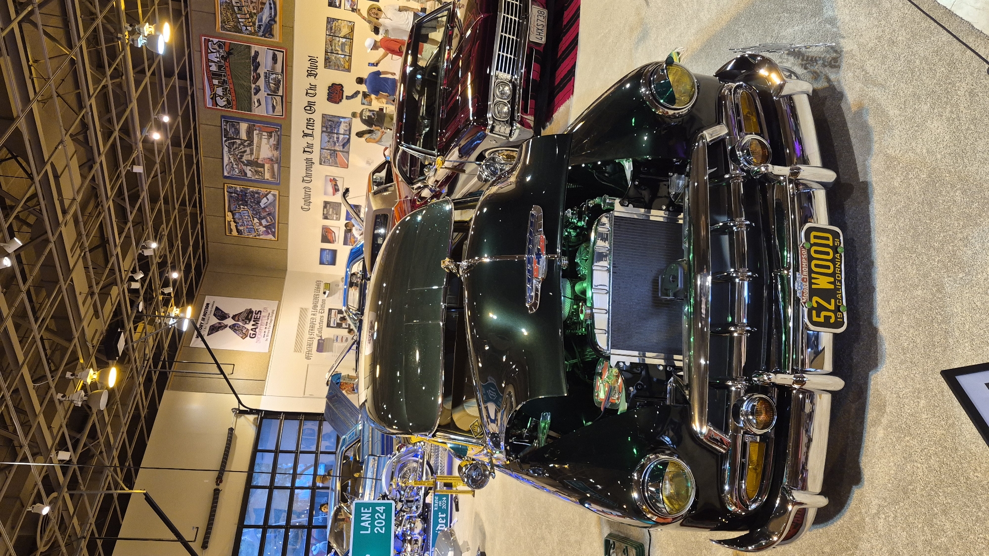 The image depicts a car show, likely indoors, with several vintage cars on display. In the foreground, a dark green 1951 Chevrolet sedan is prominently featured, its hood open, revealing a green-lit engine. The car has a chrome grille, bumper, and accents, and a yellow license plate that reads "52 WOOD 19 CALIFORNIA 51". Behind the Chevrolet, a burgundy vintage car, possibly a 1967 Chevrolet Impala, is also visible, along with other cars in the background. The environment appears to be a large hall with a high ceiling, exposed beams, and track lighting. Several framed posters adorn the wall in the background, some featuring car-related imagery and text. A few people are seen in the background, observing the cars. The flooring is a light-colored carpet. The overall atmosphere suggests an exhibition or event dedicated to classic automobiles. The specific location of the city of Sacramento, United States of America, is not explicitly indicated in the image, but the presence of a California license plate suggests it is in California.