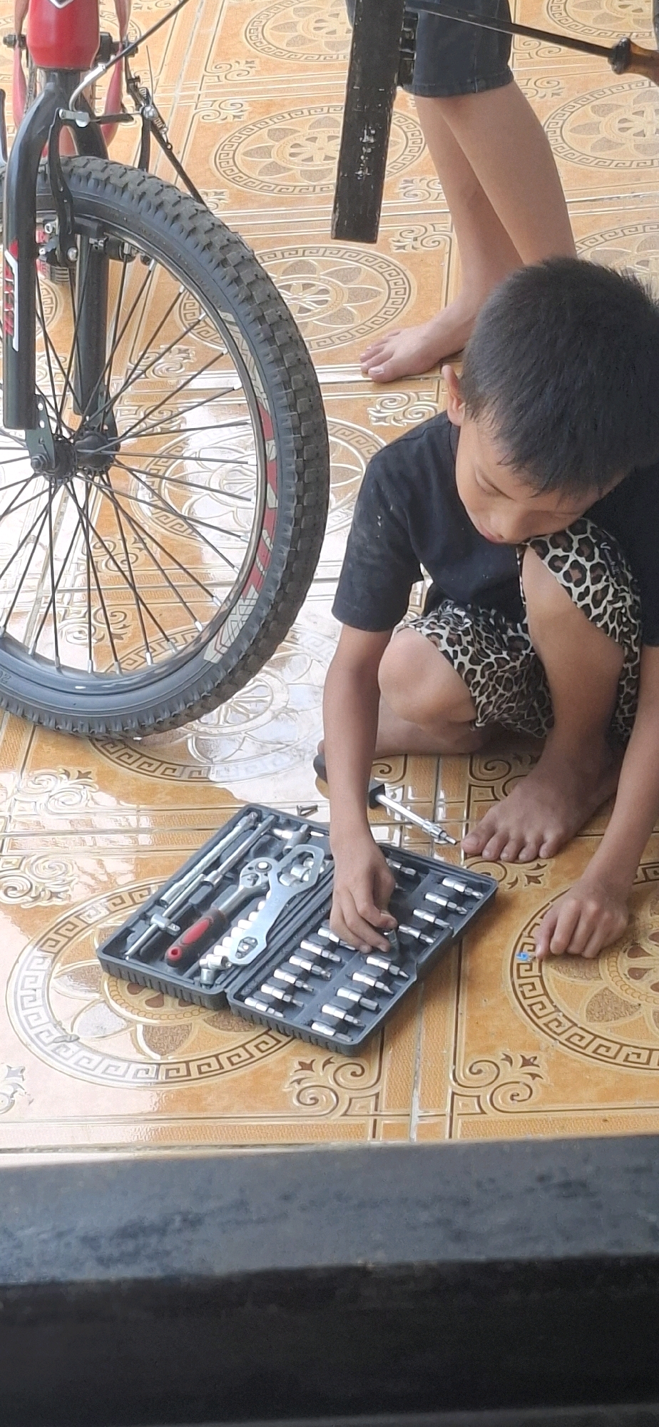 In Cibinong Hilir, Indonesia, a young boy with dark hair and wearing a black t-shirt and leopard-print shorts is kneeling on an orange tiled floor. He is focused on a grey tool case filled with various metal tools, including sockets and a wrench. His right hand is reaching into the case. To his left, a bicycle wheel with a black tire and spokes is partially visible. The floor is decorated with a repeating circular pattern. In the background, another person's legs and feet are visible, suggesting the presence of another individual. The scene appears to be outdoors, possibly a backyard or patio.