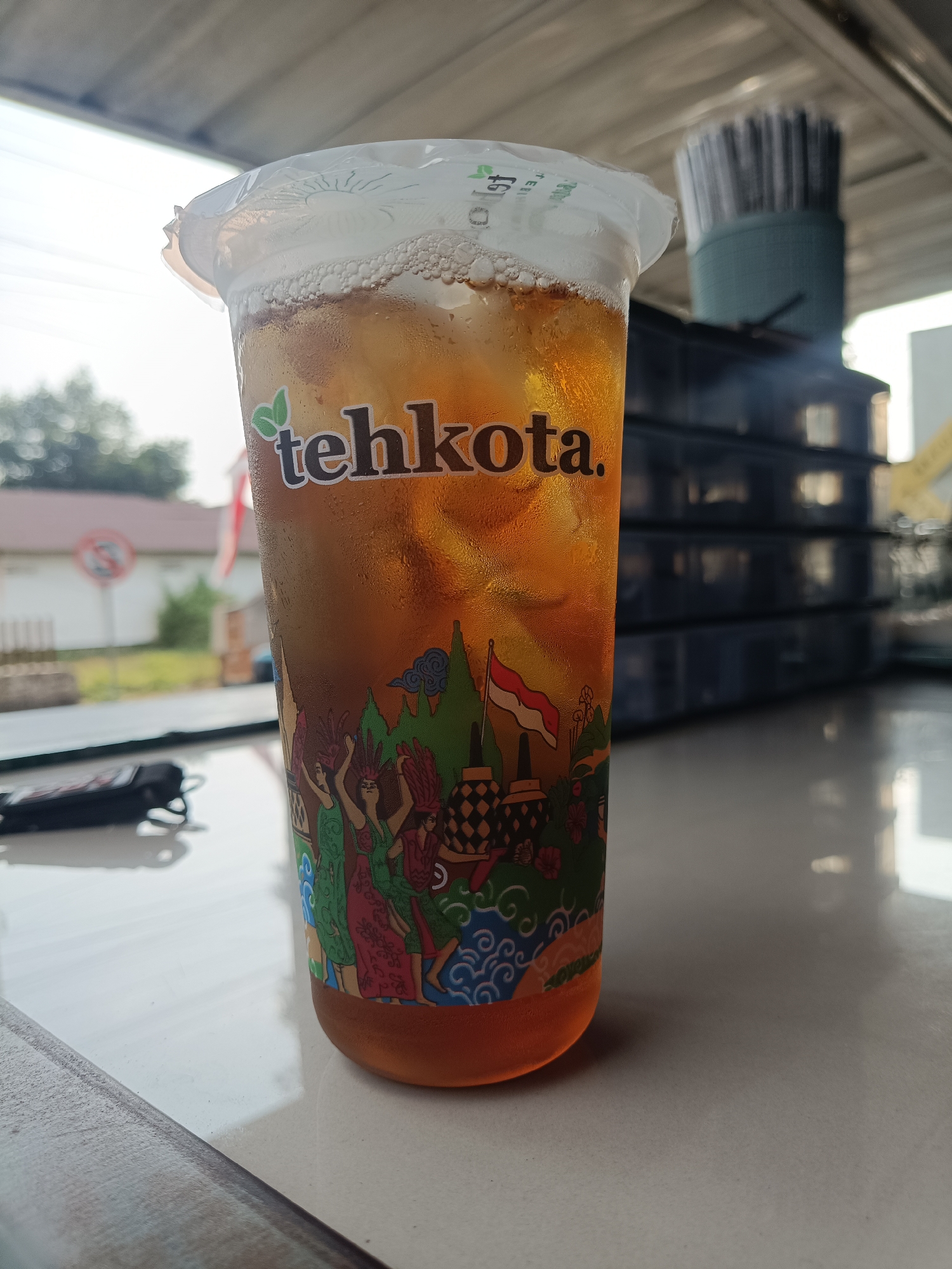 A close-up, eye-level shot features a clear plastic cup filled with iced tea. The cup is branded with the "tehkota." logo and has a decorative wrap depicting Indonesian cultural motifs, including stylized figures, buildings, and the Indonesian flag. The iced tea is amber in color and contains ice cubes and a layer of foam on top. The cup is placed on a white, reflective surface. In the background, out of focus, are dark blue stacked containers, a cylindrical object with numerous grey vertical lines, and greenery and buildings suggesting an outdoor or semi-outdoor urban setting. A black object, possibly a keychain or phone, rests on the surface to the left of the cup. The lighting suggests it is daytime, possibly morning or afternoon, with sunlight casting reflections on the surface. The image appears to have been taken in Jember, Indonesia, given the cultural imagery on the cup.