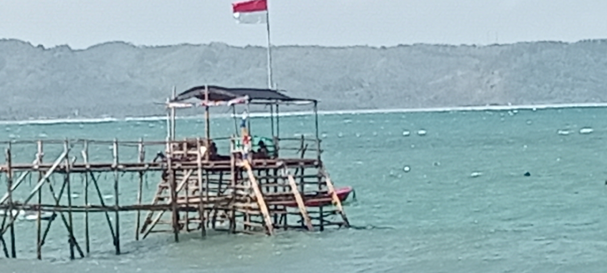A wooden pier extends into the turquoise water at Desa Pagandaran, Indonesia. The pier has a raised platform with a dark canopy and a flagpole displaying the Indonesian flag. A red dinghy is tied to the pier. The water is choppy, with ripples and some debris visible. In the background, a hazy, green mountain range borders the horizon, with a faint line of white surf breaking along the coast. The sky is overcast and a pale grey.