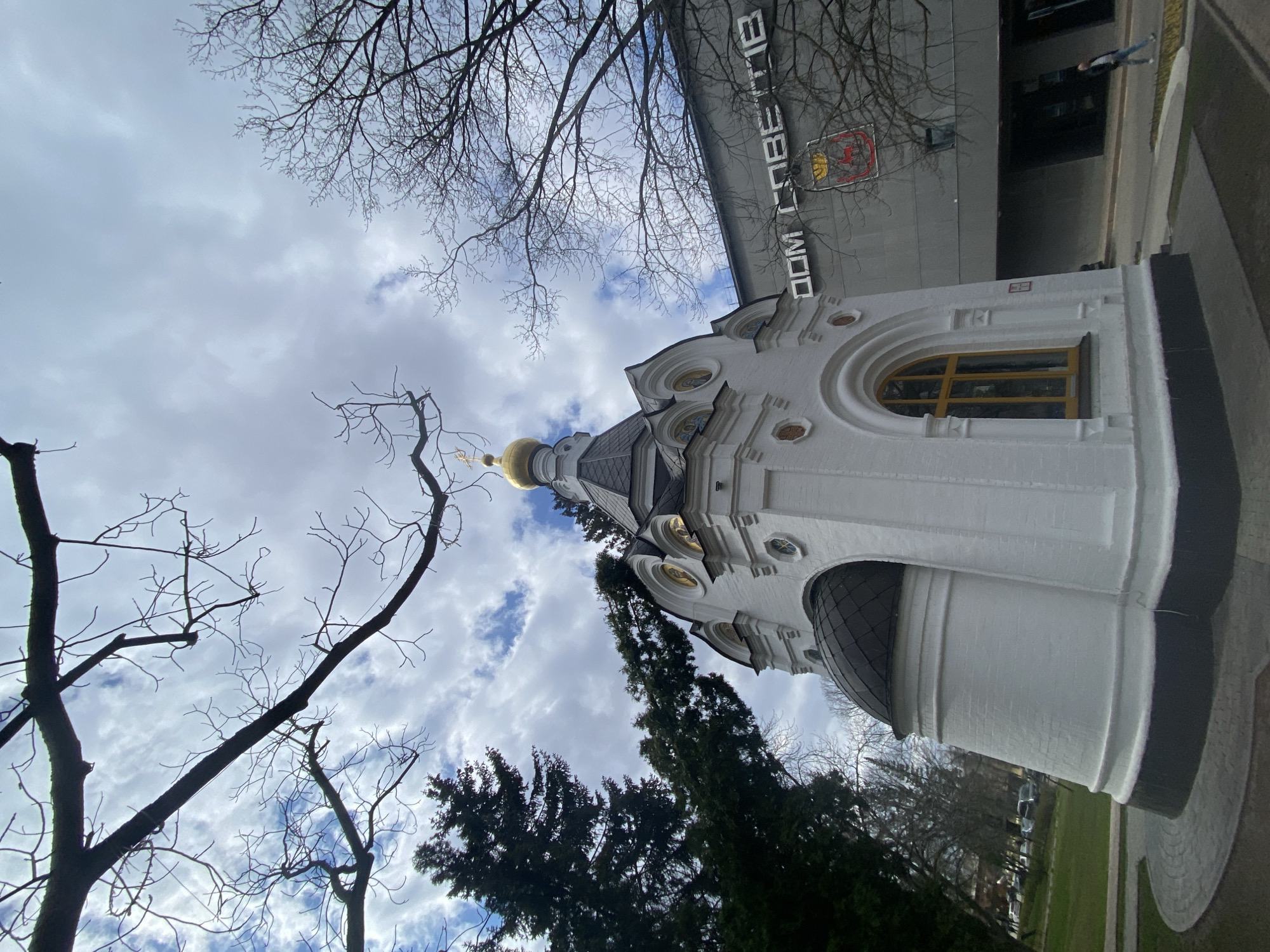 A slightly tilted, upward-looking shot captures a white, ornate architectural structure resembling a chapel or church, topped with a gleaming golden dome and smaller dark domes. The building features arched windows, some with yellow panes, and decorative pilasters. Bare tree branches, characteristic of late autumn or early spring, frame the view against a mostly cloudy sky with patches of blue.
In the background, a modern, grey building is visible, bearing Cyrillic text that reads "ДОМ СОВЕТОВ" (House of Soviets) and a red and yellow emblem. A faint figure of a person can be seen walking near this building. Dark green coniferous trees are also present in the mid-ground, and a grassy area with a paved path is visible in the lower right. The time appears to be daytime, under an overcast but bright sky. The location cues, including the church's architectural style and the Cyrillic text, align with Nizhny Novgorod, Russia.