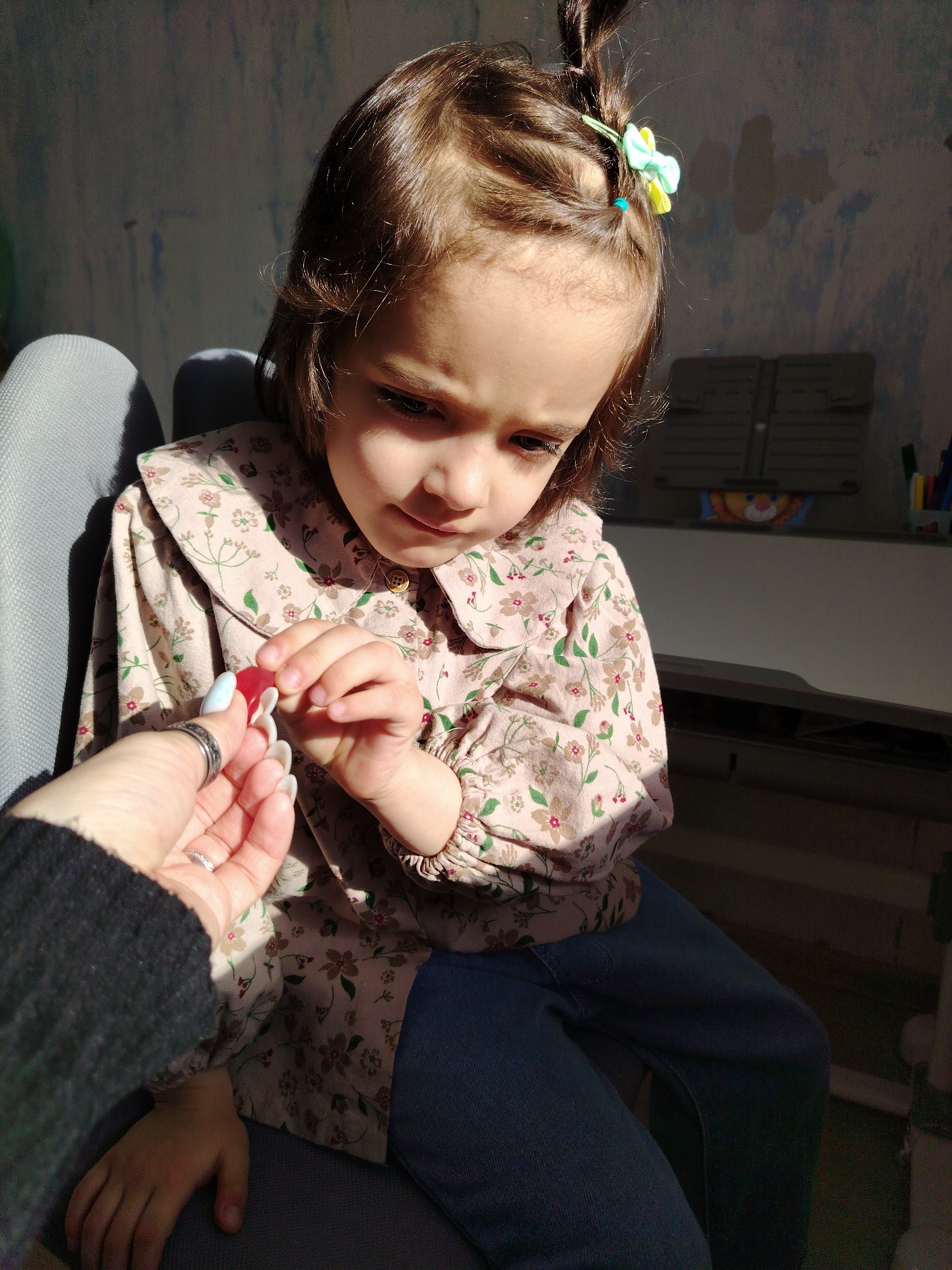 A young girl with brown hair partially styled in a high ponytail with a light green and yellow hair accessory sits on a gray chair and looks at a red candy held by an adult’s hand. The girl is wearing a long-sleeved beige shirt with a floral pattern and blue jeans. The adult's hand has long painted fingernails and is wearing a silver ring. The background features a partially visible white desk and a blue wall with paint imperfections. The scene is illuminated by strong sunlight. The location is not discernible from the image.