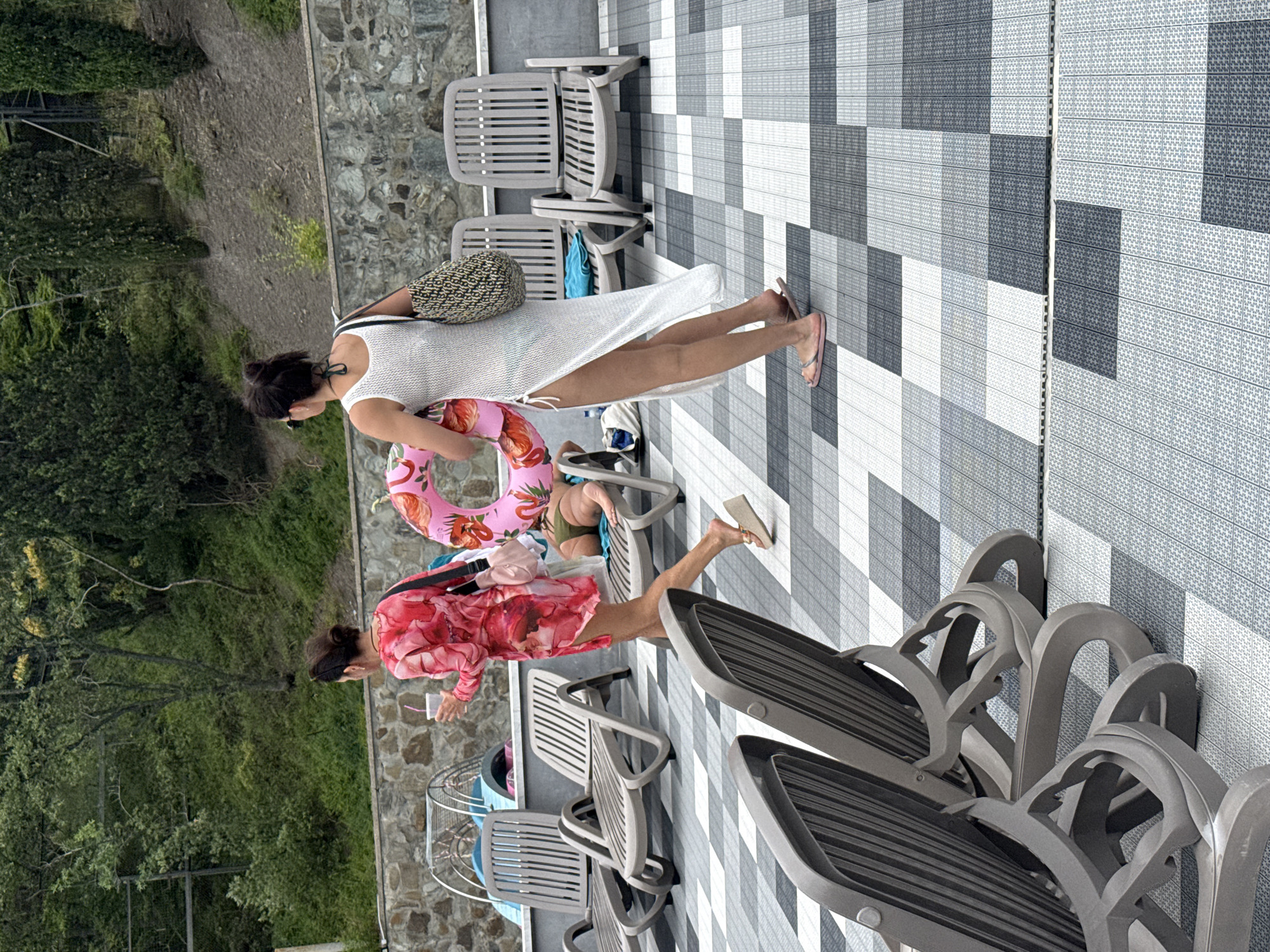 The image depicts a poolside area with two women and several lounge chairs. The foreground is dominated by the tiled ground and several empty, grey plastic lounge chairs. In the mid-ground, one woman, wearing a red floral dress and holding a drink, walks away from the camera, carrying a pink bag over her shoulder. She is walking past a man who is lying on a lounge chair wearing green swim trunks. To her right, another woman walks towards the camera wearing a white sheer cover-up dress and carrying a pink inflatable ring with a flamingo pattern. She has a large patterned tote bag over her shoulder. Behind the people, a stone wall forms the boundary of the pool area, with lush green foliage and trees visible on the hillside beyond. The overall setting suggests a warm weather resort or public pool area.