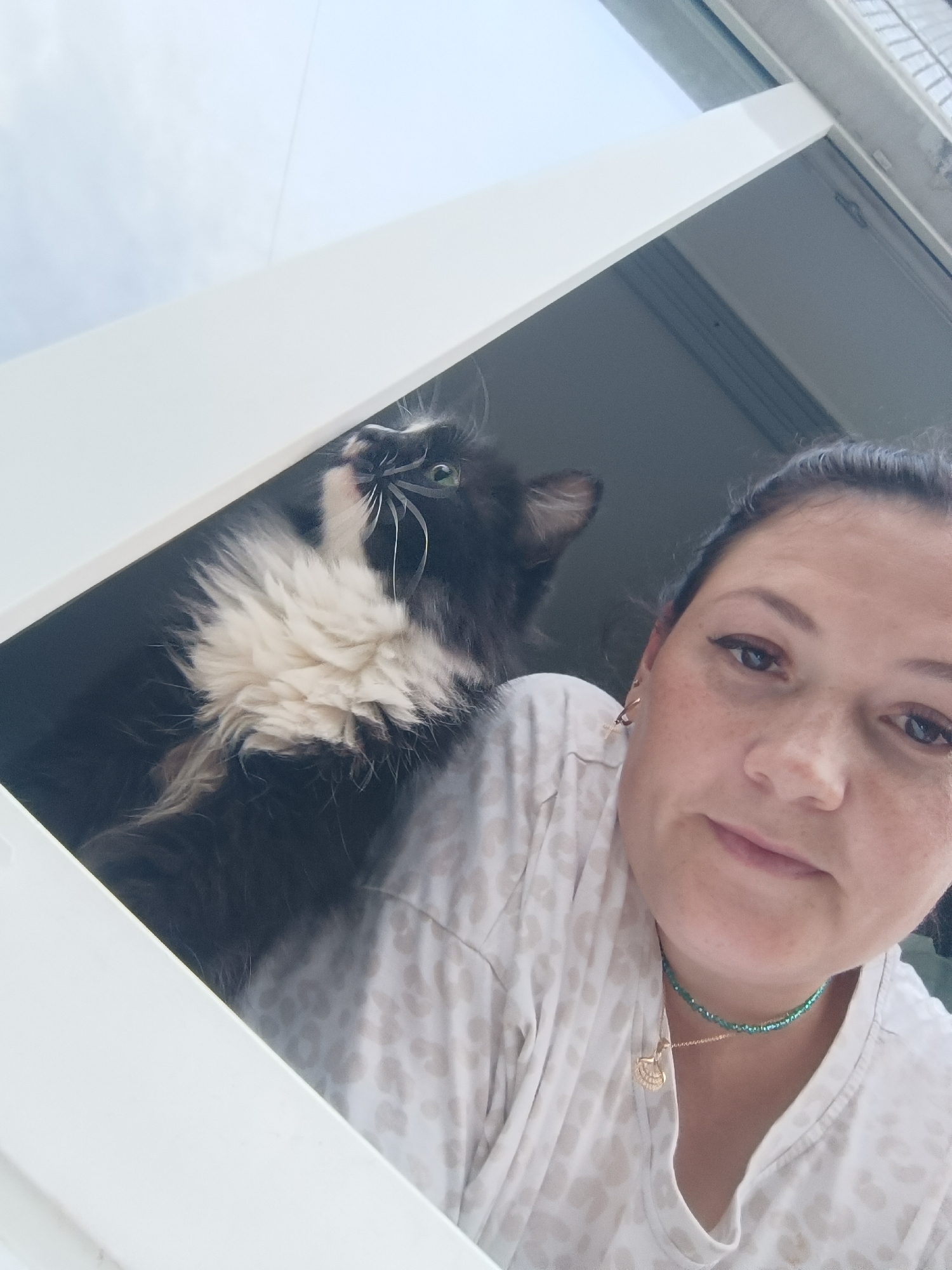 This is a selfie taken from a low angle of a woman and a cat in a window. The woman is on the right side of the frame, looking towards the camera. She has dark hair pulled back, and is wearing a light-colored, patterned shirt. Around her neck is a green beaded choker and a gold pendant necklace. She has freckles on her face and is wearing eyeliner. To her left is a black and white fluffy cat. The cat is looking upwards, away from the camera, with bright green eyes and long white whiskers. The cat has a thick white ruff around its neck and chest. They are both positioned within a white window frame, with a view of a cloudy sky. The scene appears to be outdoors, possibly on a balcony or in a window overlooking an urban or suburban area. The location is Chaikovsky, Russia.