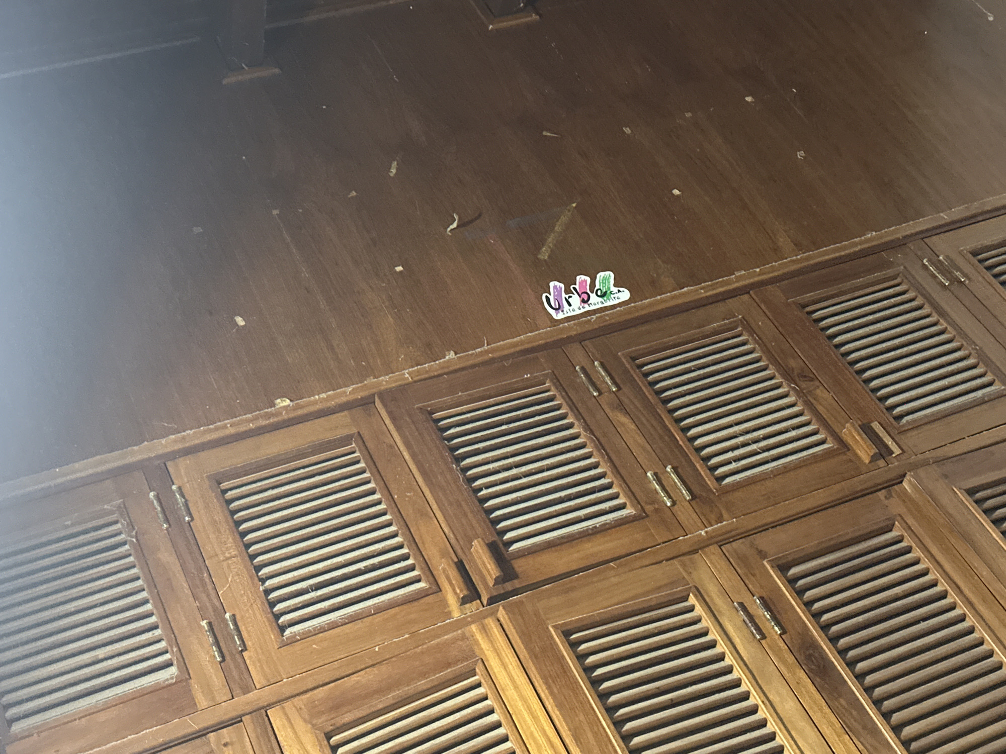 The image displays an indoor scene in Juan Griego, Venezuela, captured from a tilted, high-angle perspective. The upper section of the frame features a large, horizontally oriented, light-brown wooden panel exhibiting a slightly distressed texture and scattered light-colored debris. A soft, bright light source illuminates the upper-left quadrant. Positioned centrally on this upper panel, slightly above the lower wooden structures, is a white rectangular sticker with colorful lettering that reads "urbo C.A." and "Isla de Margarita" below it. Below the main wooden panel, a series of interconnected, louvered wooden cabinet doors or panels occupies the lower and mid-sections of the image. These doors are constructed from a similar light-brown wood and are fitted with brass-colored hinges. Visible dust accumulation is present within the louvers and along the seams of these wooden structures. No individuals or dynamic actions are depicted.
