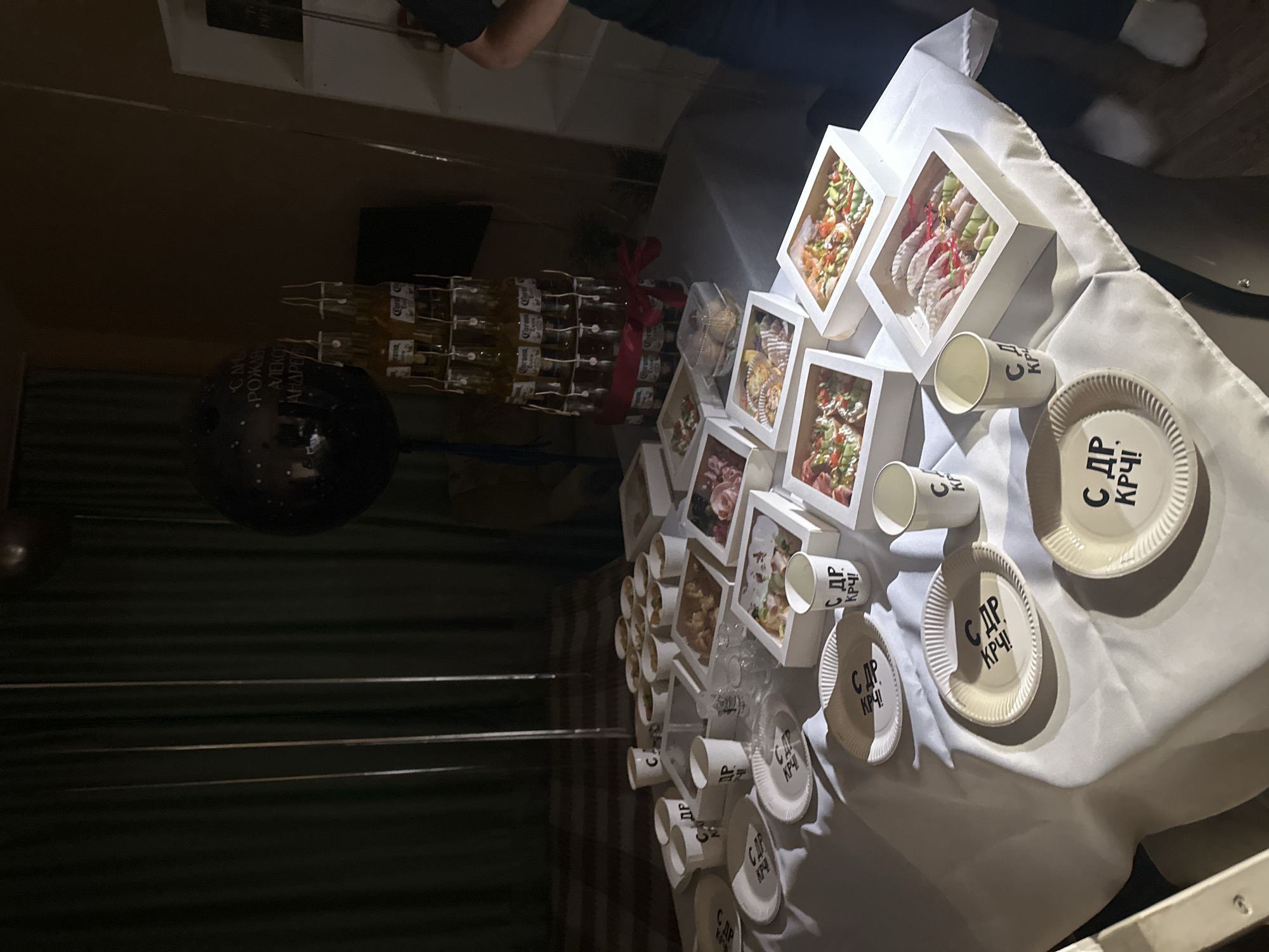 This is a medium shot, eye-level, indoor photograph featuring a food spread on a table covered with a white tablecloth. The focus is on the food and party supplies arranged for what appears to be a celebration. In the background, dark green curtains hang, and a large black balloon with silver speckles is visible, with text that is partially obscured but appears to say "С ДР" (Happy Birthday) and a name that is difficult to decipher.
Dominating the center of the table is a pyramid-like structure constructed from multiple bottles of Corona Extra beer. A red ribbon is tied around the base of the beer bottle tower.
Scattered across the table are white paper plates and cups, many of which are printed with the Cyrillic text "С ДР КРЧ!". Several white square boxes contain various food items, appearing to be appetizers or small meals, with visible ingredients like seafood, vegetables, and possibly meats. There are also smaller white cups and a few clear plastic containers with lids. A few glasses are also present.
The lighting in the scene is dim, casting shadows and creating a somewhat moody atmosphere. The overall presentation suggests a casual party or gathering. There are no identifiable landmarks or clues within the image to determine a specific location within the city of Perm, Russia. The image does not contain any discernible individuals.
