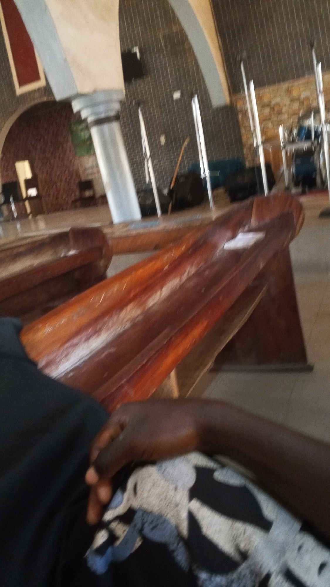An interior shot shows pews in a place of worship. The pews are made of polished dark wood and appear to be empty. The background shows a patterned wall, a large pillar, and some equipment that may be related to sound or music. A person's arm and hand are visible in the foreground, resting on a patterned fabric. The lighting suggests it is daytime, though the overall atmosphere is dim.
