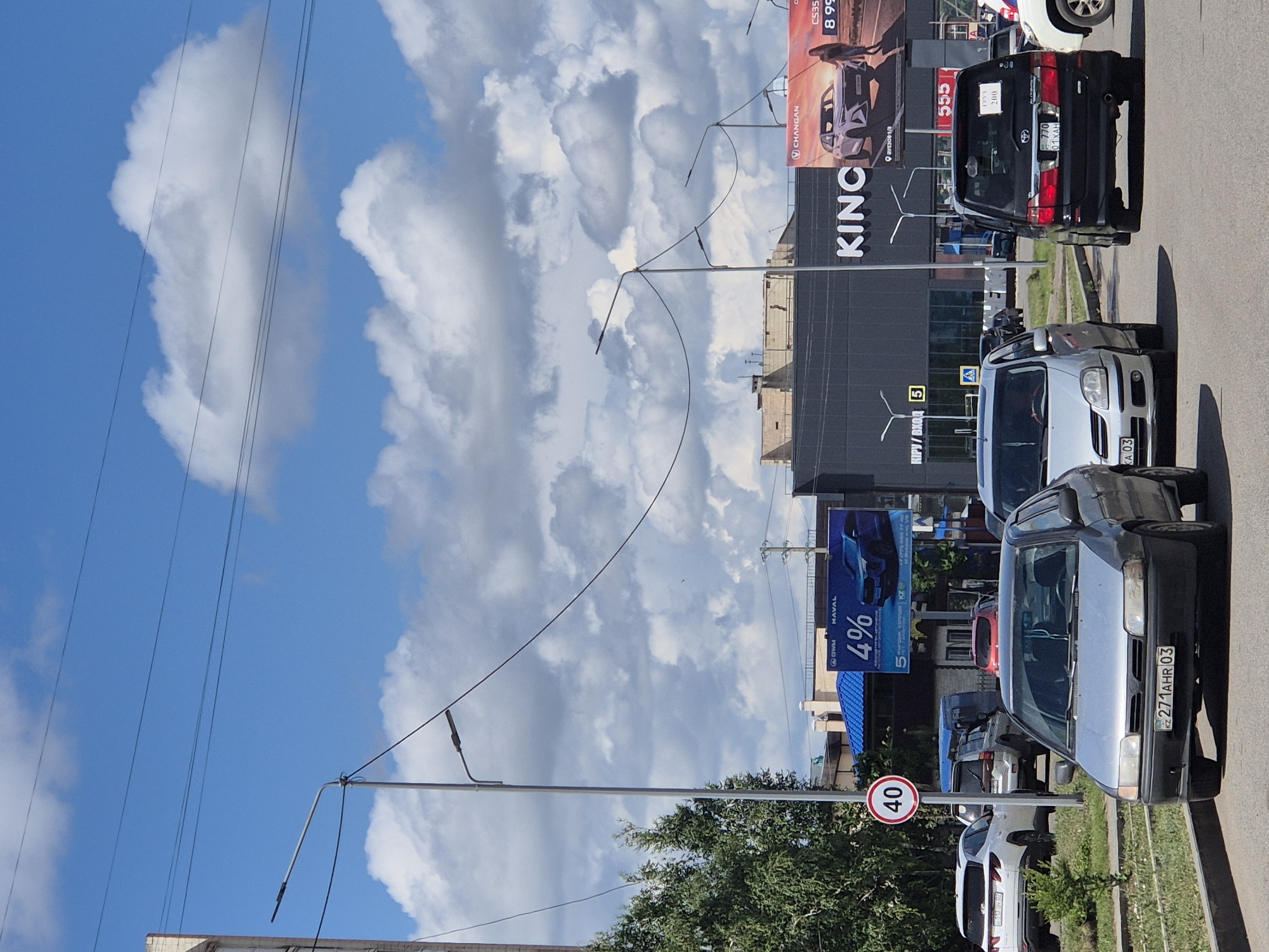 The image depicts a street scene in Kokshetau, Kazakhstan, under a bright, cloudy sky. In the foreground, several cars are visible on a paved road. A silver sedan is prominent, with license plate KZ 271 AHR 03. To its right is a black minivan with license plate KZ 770 01 XAH. A white SUV is parked on the left side of the road, and another white SUV is partially visible in the background.
Further down the street, a large, dark building with "KING" prominently displayed on its facade is visible. This building appears to be a commercial establishment. To the left of this building, a blue structure with a distinctive roof is present. Advertising billboards are displayed on the side of the "KING" building and elsewhere in the background. One billboard features an advertisement for "CHANAN" and "HAVAL" with a "4%" offer. Another billboard displays the number "555".
Streetlights and power lines are visible throughout the scene, indicating an urban environment. A speed limit sign showing "40" is mounted on a pole near the left side of the road. Trees and greenery line the side of the road, suggesting the presence of a sidewalk and possibly a park or residential area. The overall impression is a typical day in a moderately busy urban area.