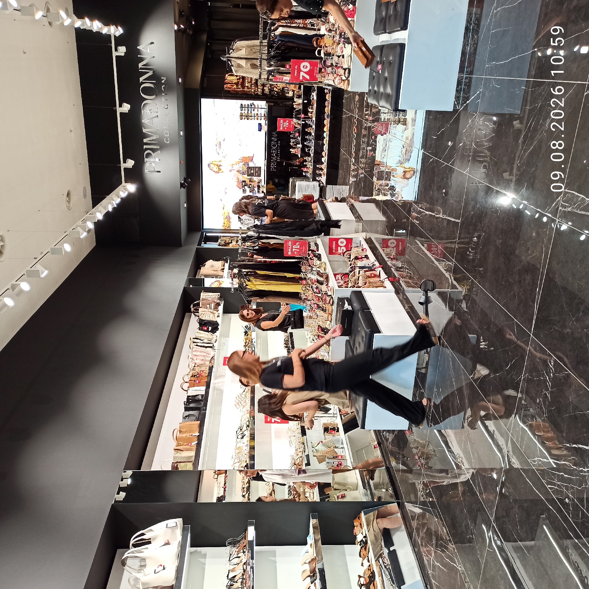 This is a photograph taken inside a Primadonna Collection store, likely in Molfetta, Italy, as indicated by the date stamp. The scene depicts a retail environment focused on women's fashion, specifically shoes and handbags.
The store features sleek, modern design with dark, reflective marble flooring that mirrors the displays and shoppers. The walls are dark, with white shelves and displays showcasing a variety of merchandise. Overhead, modern track lighting illuminates the space.
In the foreground, a woman in a black t-shirt and black pants is walking from left to right across the frame. She appears to be a shopper or employee. She is partially obscured by the frame. Another woman, also in black, is visible further in the background, standing behind a rack of clothing. To her left, a woman in a white dress is looking at items on a shelf.
Various displays are visible:
* On the left, shelves hold rows of women's shoes.
* To the right of that, a wall display showcases a collection of handbags.
* Further to the right, racks of clothing are displayed, with some items on hangers.
* In front of the clothing racks and further right, shelves are laden with more shoes and handbags.
* Some displays have signage indicating sales, such as "SALDI -70%" (Sales -70%) and "BORSE 50" (Bags 50).
Reflections on the floor provide glimpses of other shoppers and displays, adding depth to the scene. A large digital screen displaying an advertisement or fashion imagery is visible in the background behind the clothing racks. The overall impression is of a well-organized and busy fashion retail store. The date stamp "09.08.2026 10:59" suggests the photograph was taken on August 9, 2026, at 10:59 AM.