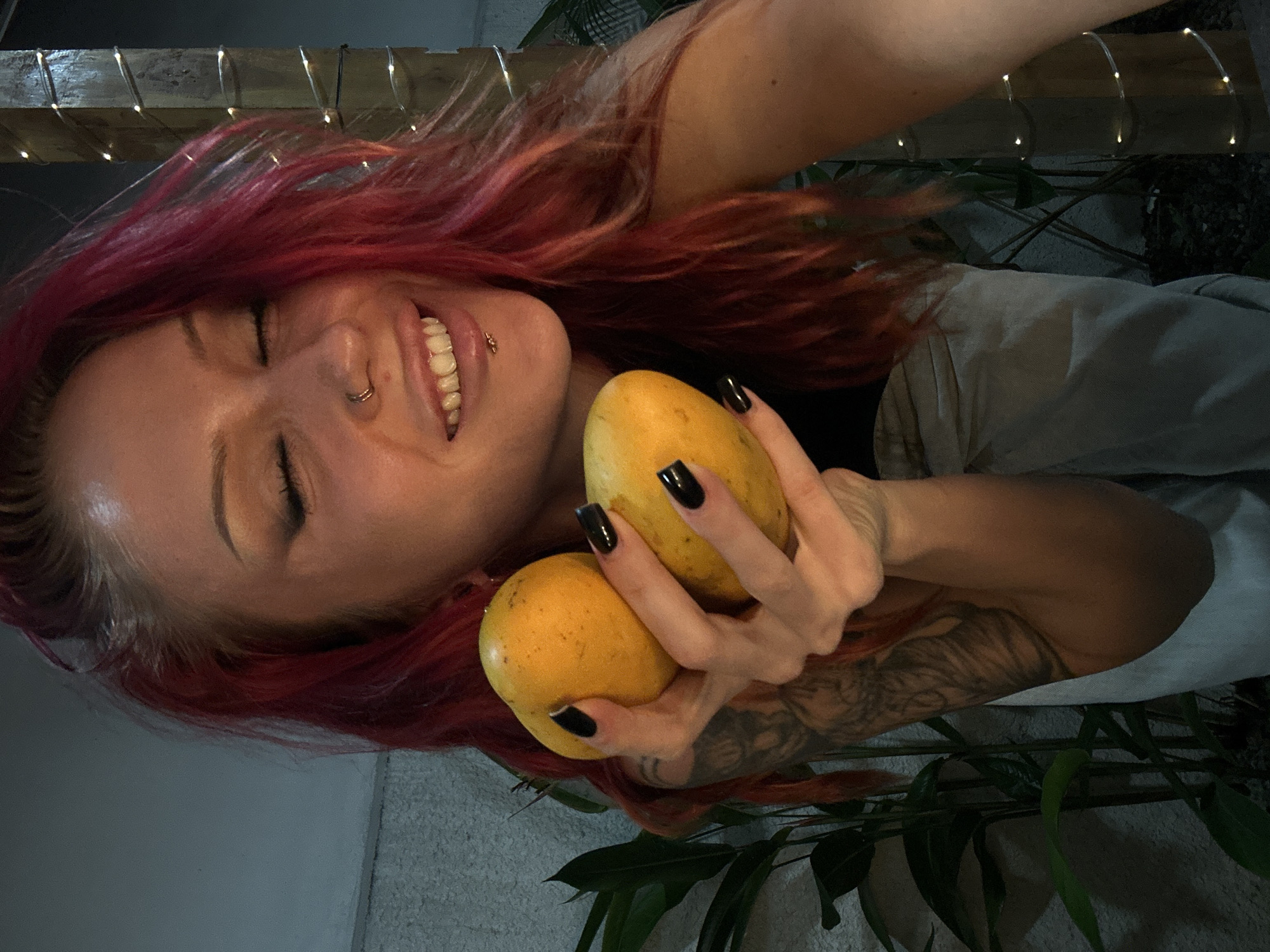 A woman with red hair and dark fingernails is holding two mangoes. She is wearing a black top and light-colored pants. She has a nose ring and a lip piercing. She is standing in front of a wall with plants and a wooden post decorated with lights.