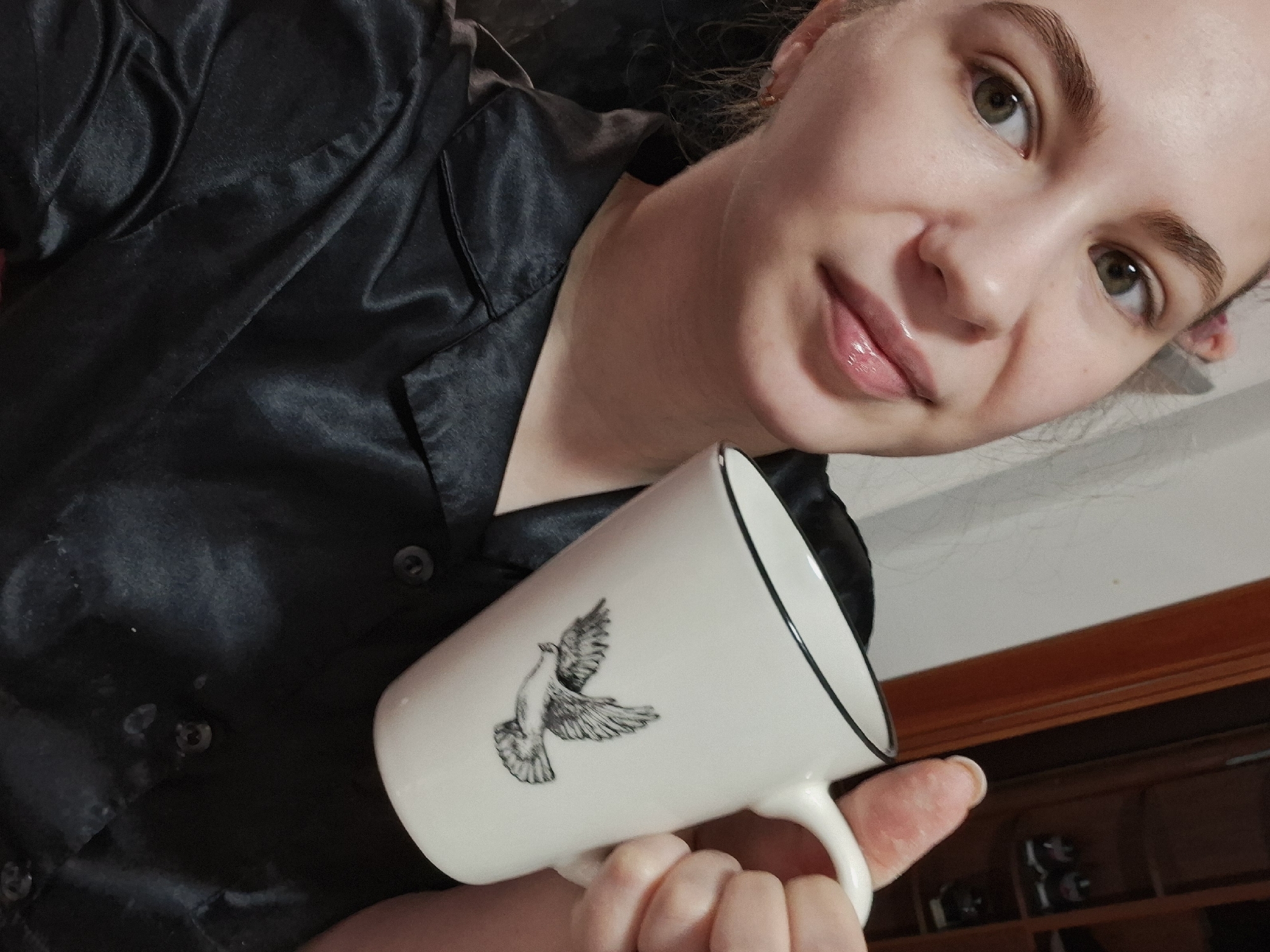 A woman with fair skin and light brown hair pulled back is holding a white mug with a black-and-white drawing of a dove on it. She is wearing a black satin shirt with buttons. The background shows a wooden cabinet. The location is not identifiable as Chaikovsky, Russia, based on visual cues in the image.
