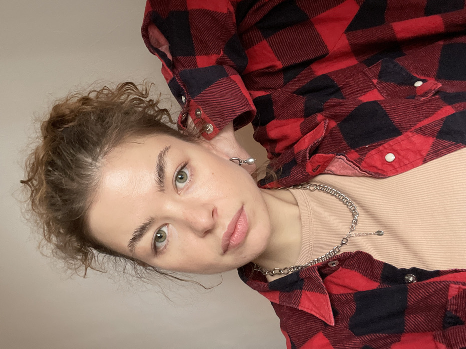 A close-up portrait features a female subject indoors. She has a fair complexion, green eyes, and light brown, curly hair secured upwards. Her expression is neutral, with her gaze directed towards the camera. She is attired in an open red and black plaid collared shirt over a beige ribbed top. A silver-toned chain necklace with a small drop pendant is visible around her neck. A silver-toned hoop earring featuring a white pearl drop adorns her left earlobe. Her right arm is bent, with the hand positioned behind her head, partially obscured by her hair. The background is a plain, light-colored wall. There are no visual cues within the image to identify the specific location as Boreti, Montenegro, or any other city.
