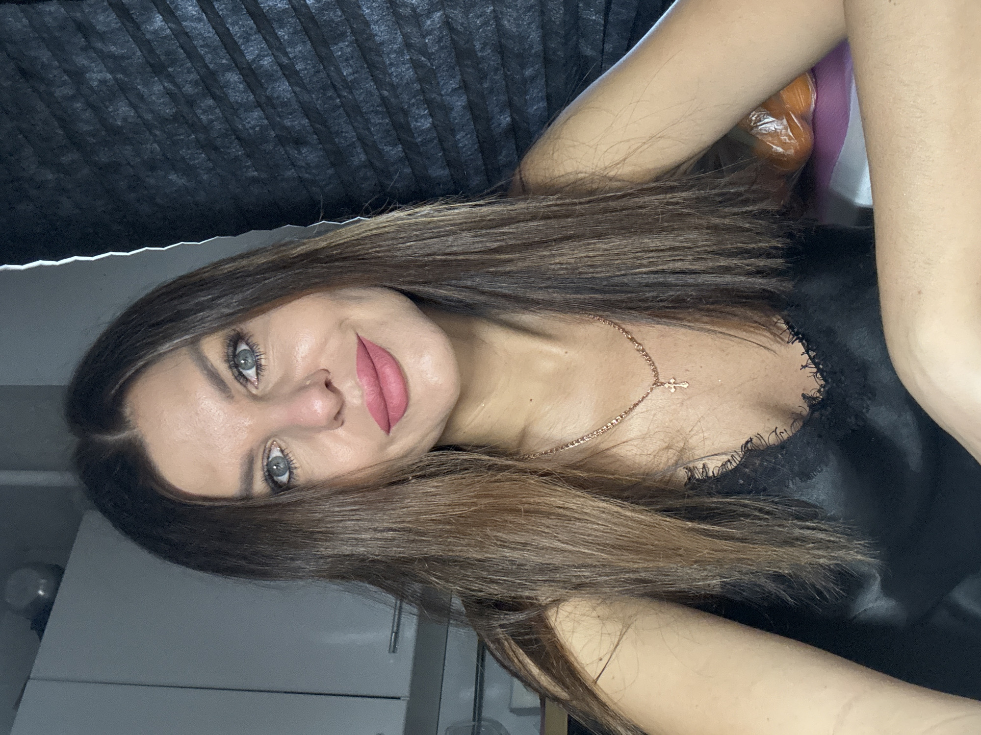 A young woman with brown hair and blue eyes is positioned with her head slightly tilted to the left. She wears pink lipstick and a delicate gold necklace with a small cross pendant. She is wearing a black top with lace trim. Behind her, a dark, pleated material hangs like a curtain or blind. The background also shows a white wall and what appears to be a piece of furniture, such as a cabinet. The location is potentially in Chaikovsky, Russia.