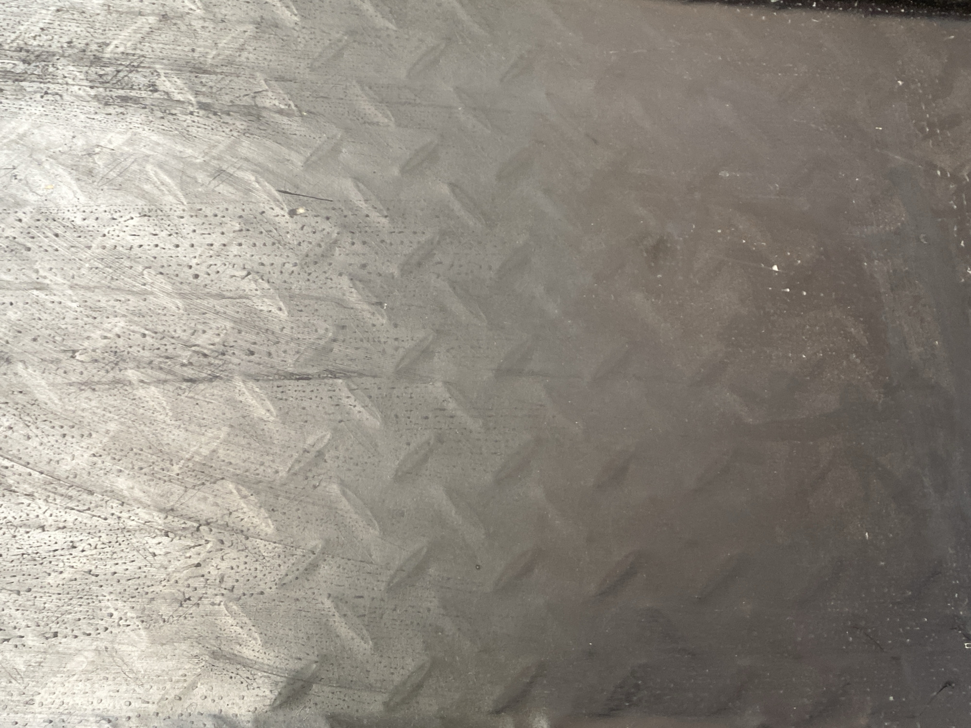 The image shows a close-up, full-frame view of a weathered, textured metal surface. The surface has a diamond plate pattern visible, particularly in the upper and mid-sections of the frame, where the raised ridges are more distinct. The metal appears to be gray or dark gray in color. There are numerous small water droplets or condensation marks scattered across the surface, especially concentrated in the upper half. Streaks and smudges are also present, suggesting it has been exposed to the elements and possibly handled. The lighting creates a gradient, with the top portion appearing brighter and more detailed due to the texture and droplets, while the lower portion becomes darker and smoother, with less visible texture. There are no people, animals, or specific landmarks identifiable in this abstract view of the metal surface. The image does not contain any information about its location in Jauro Yinu, Nigeria.