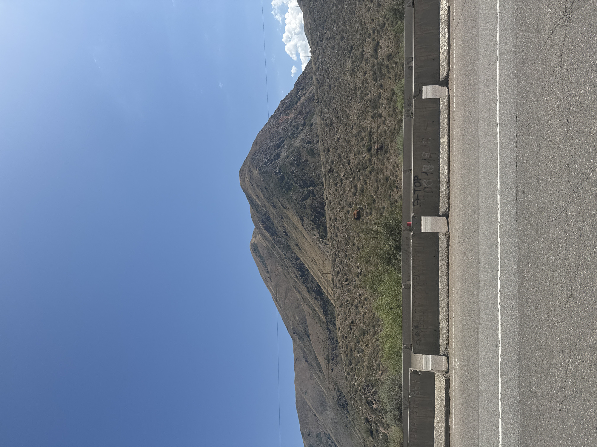 The image shows a mountain landscape under a clear blue sky. In the foreground, there is a paved road with a white line marking the lane. Concrete barriers, some of which appear to be graffitied, separate the road from the embankment. The embankment is covered in dry brush and sparse vegetation, leading up to a large, barren mountain. The mountain has visible layers and textures, suggesting a rocky, arid terrain. The location is identified as Cholok, Kyrgyzstan. The scene appears to be a roadside view of a mountainous region.