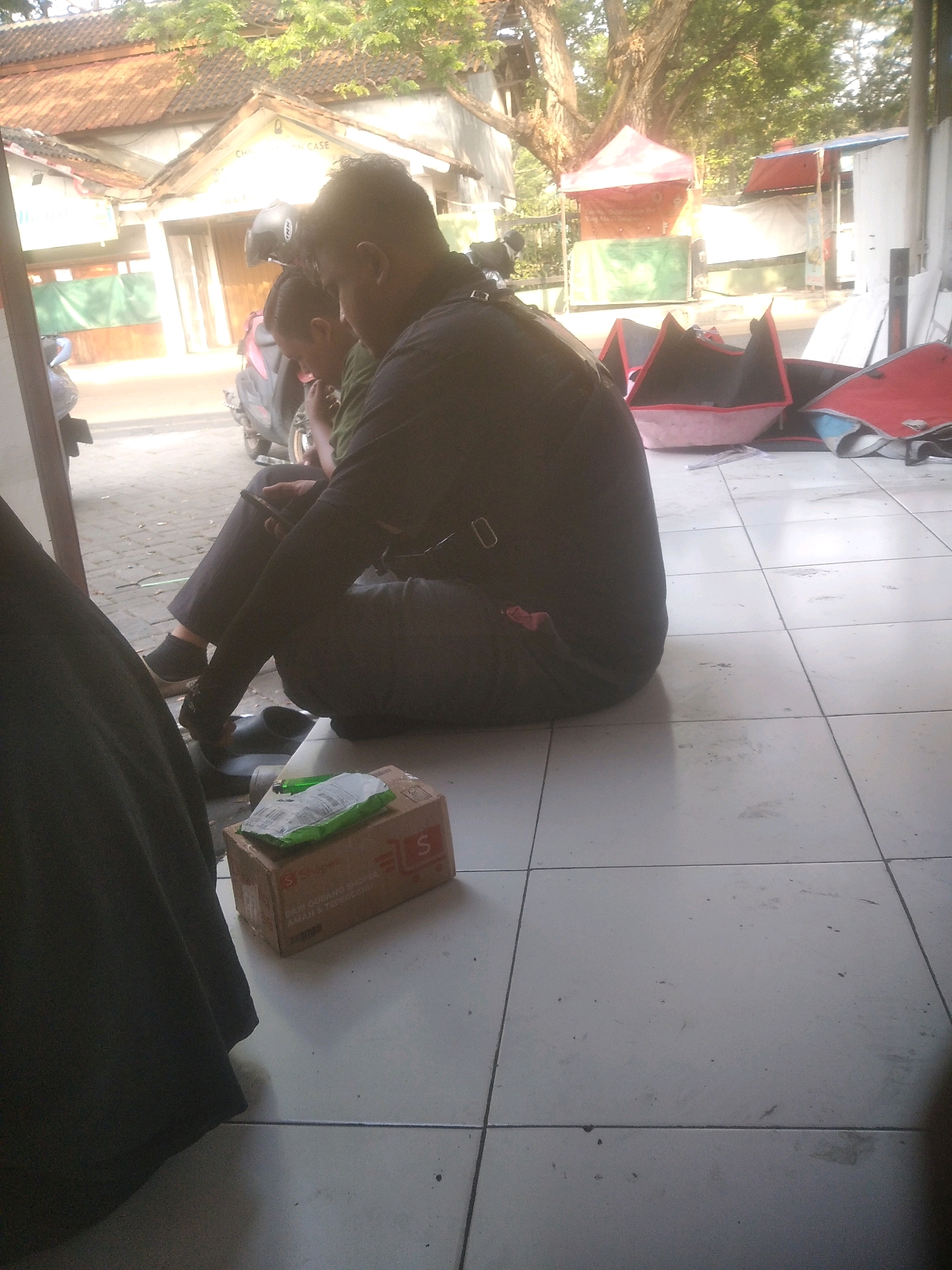 The image depicts two individuals sitting on a tiled floor in what appears to be a commercial area in Cilegon, Indonesia. The person in the foreground, on the right, is wearing a dark shirt and pants, and is looking down, possibly at a phone. Beside them, slightly to the left, is another person wearing a dark shirt and pants, also looking down. In front of the seated individuals is a cardboard box with a green package on top. A motorcycle helmet is visible near the top of the frame, resting on a motorcycle behind the seated figures. In the background, there are buildings with tiled roofs, some stalls with tarpaulin coverings, and trees. The overall scene suggests a casual waiting area or a rest stop in a bustling urban environment.