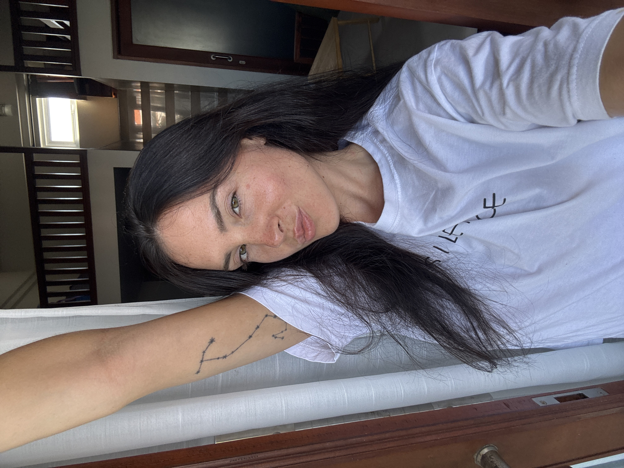 A young woman with long, dark hair and fair skin stands with her left arm raised, her hand resting near the top of the frame. She is wearing a white t-shirt with black text on it that reads "SILENCE." She is looking at the camera and puckering her lips. A tattoo of a constellation is visible on her left arm. The background appears to be an indoor space with wooden accents, possibly a mezzanine or upper level with a railing. There is a window with bright light coming through it, and a doorway on the right side of the frame. The overall setting suggests a residential or hotel room in North Kuta, Indonesia.