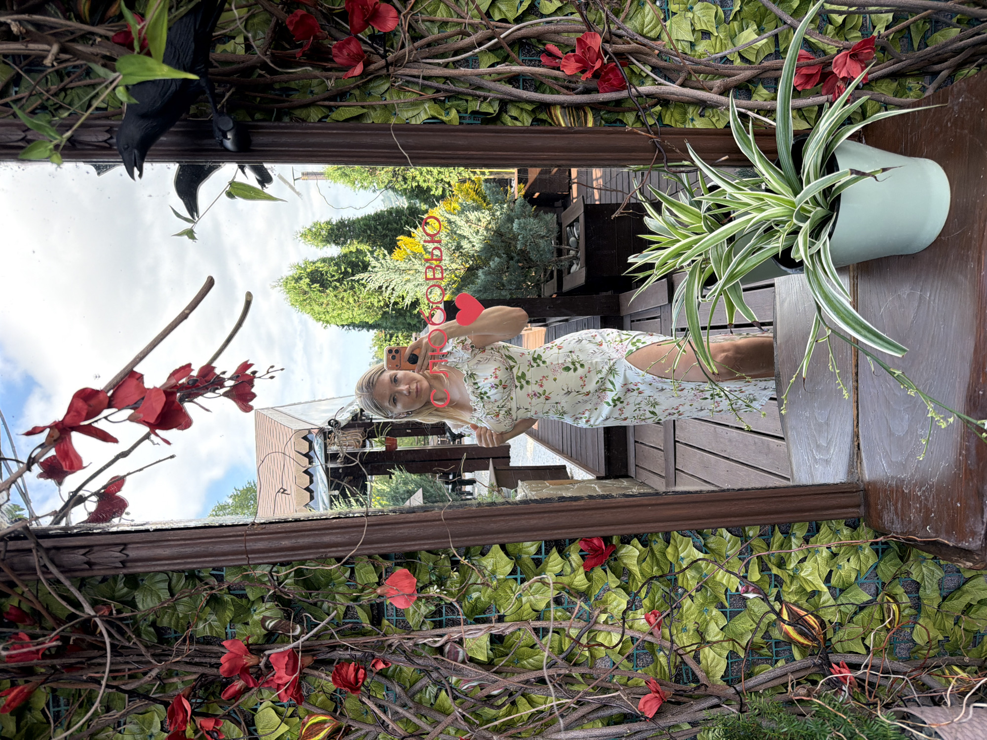 A full-body mirror selfie shows a blonde woman with fair skin and blue eyes standing in front of a decorative wall with artificial greenery and red flowers. She is wearing a light-colored, floral-print dress with a thigh-high slit and a ruffled neckline. She is holding a light orange smartphone in her right hand, positioned to take the photo. The mirror reflects a wooded area with trees, a building with a brown roof, and a cloudy sky. Two black crow figures are perched on the frame of the mirror. In the foreground, a light green pot contains a spider plant. The scene is outdoors, possibly on a patio or balcony, given the wooden decking and potted plants. The woman is in the city of Nizhnyaya Shilovka, Russia.