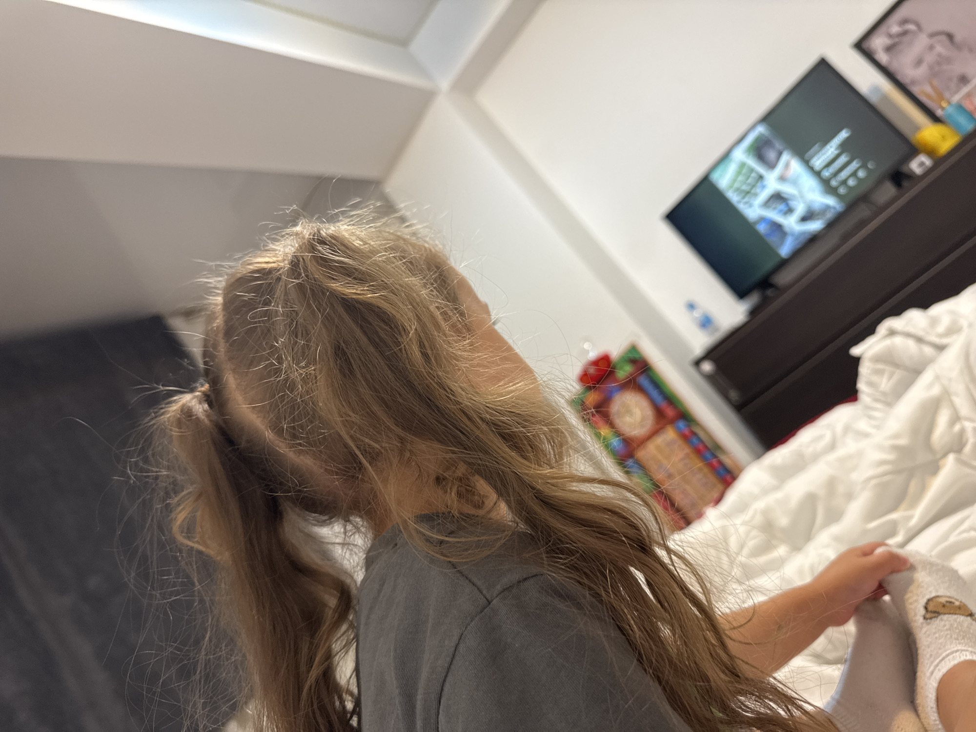 The image shows a close-up, from behind, of a young child with long, blonde, pigtail braids. The child is wearing a dark gray t-shirt and white socks with a bear design. The child is lying on a white, rumpled bed. To the right of the bed is a dark brown dresser, on top of which sits a flat-screen television displaying images. Behind the dresser, a framed picture can be seen. Further to the right, a colorful item with a patterned design is visible. The room appears to be a bedroom. The lighting suggests it is daytime, possibly late afternoon or early evening, with light coming from an unseen source, illuminating the ceiling and the bed. The scene is indoors, likely in a home. The child's posture suggests they are relaxed, possibly watching the television or resting. There is no discernible weather information. There is no visible text in the image.