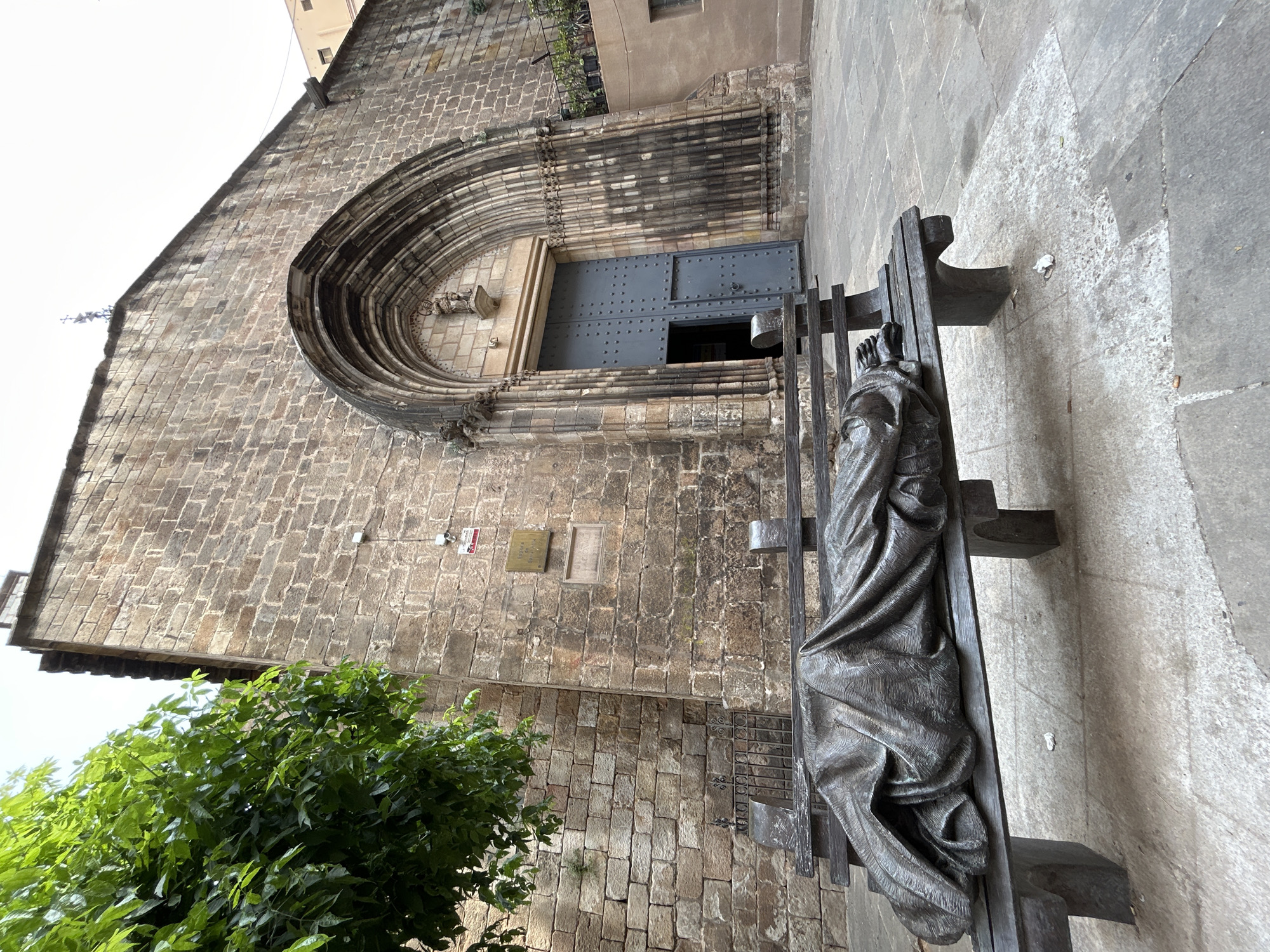 A daylight photograph captures the exterior of a historic stone church, likely the Church of Sant Felip Neri, in Barcelona. The scene is dominated by the ancient facade of the building, constructed from irregular blocks of light brown and grey stone. A large, deeply recessed arched doorway forms the central entrance, featuring multiple concentric decorative bands. Within this arch, dark grey metal double doors are visible, with the right door slightly ajar, revealing a dark interior. A small sculpted figure is nestled in a niche above the doors.
To the right and slightly in front of the church entrance, a dark bronze statue rests horizontally on a simple, dark metal bench or platform. The statue depicts a draped human figure, its form largely obscured by voluminous folds of fabric, with only its bare feet distinctly visible at one end. The overall impression is one of solemnity.
On the stone wall to the left of the entrance, a metal plaque is affixed, bearing the faint inscription "AÑO 1791" and text that appears to be "SANT F...", likely referencing "Sant Felip Neri". A security camera is also visible near the plaque. Lush green foliage partially obscures the lower left portion of the building. The ground is paved with light-colored flagstones. The bright sky suggests daytime conditions.
