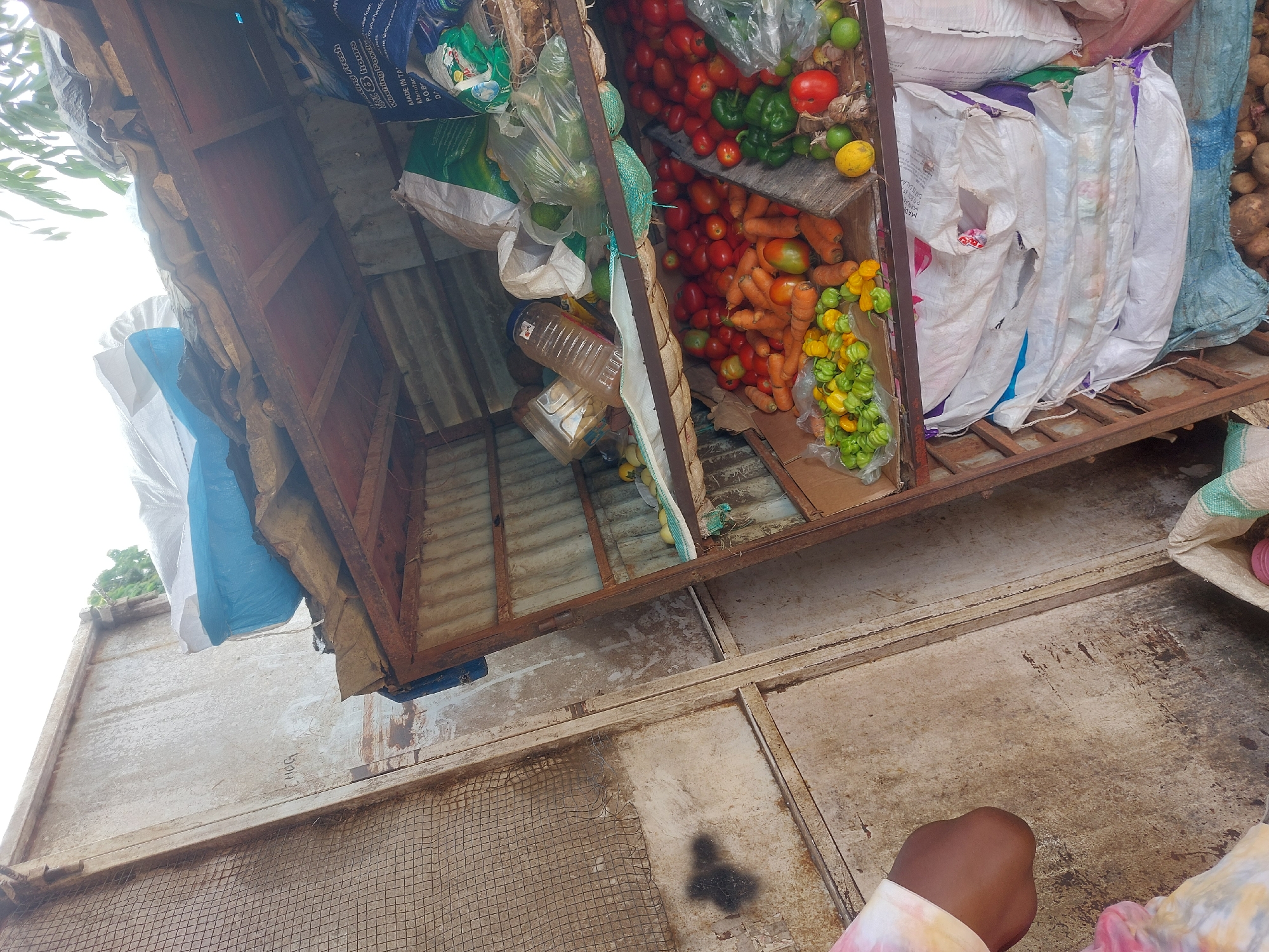 The image shows a roadside vegetable stand in Dar es-Salaam, Tanzania. The stand is constructed from metal and wood and is filled with various vegetables and bags of produce. Tomatoes, green peppers, limes, carrots, and yellow and green peppers are visible. Bags of produce are stacked next to the vegetables. A person with dark skin and a light-colored, tie-dye shirt is partially visible in the lower right corner of the image. The stand is situated on a concrete surface and a metal mesh is also visible.