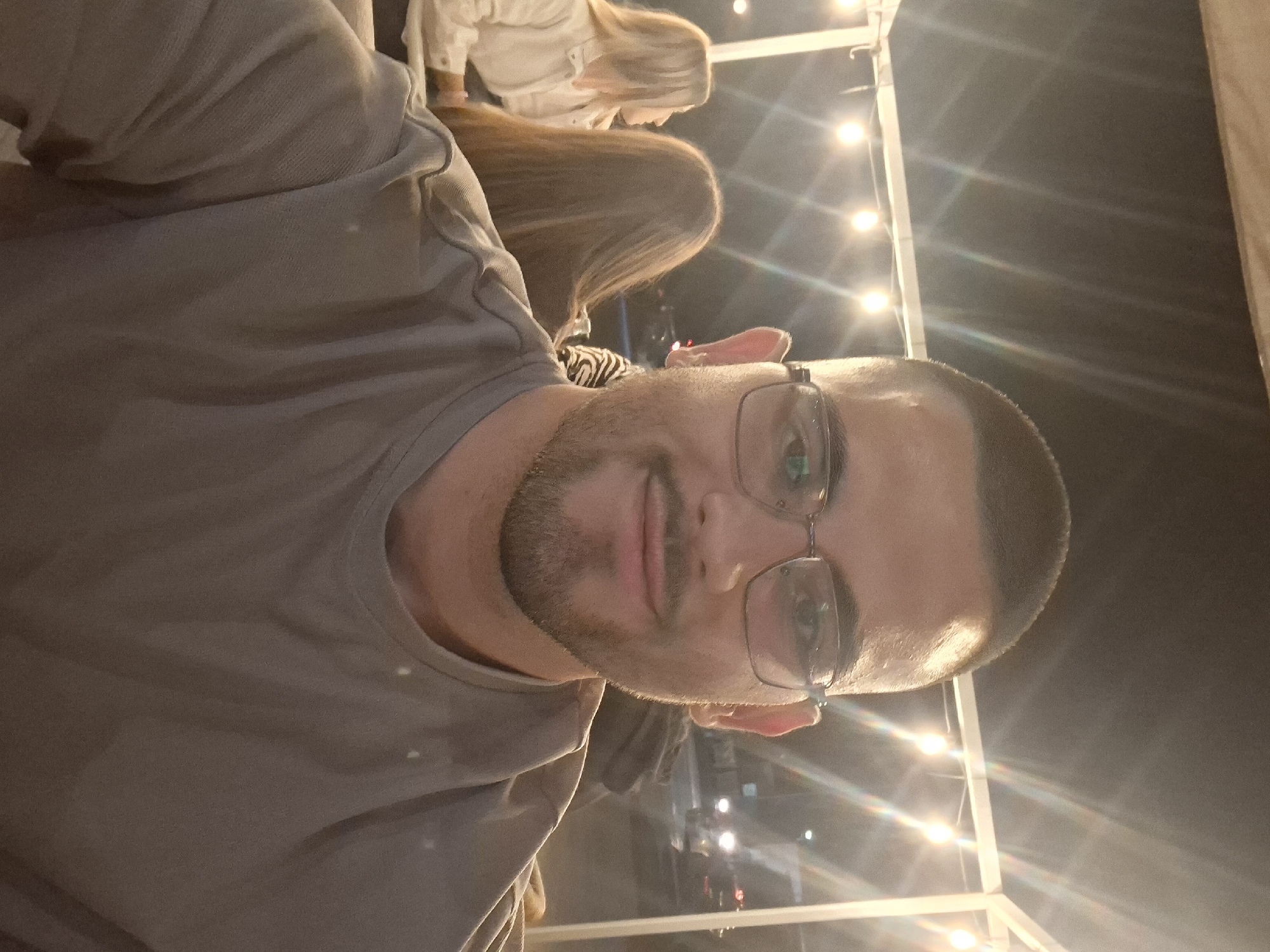 A low-angle, outdoor selfie of a man with short brown hair, a trimmed beard, and glasses. He wears a gray t-shirt. Behind him are at least two other individuals, one with long brown hair and another with long blonde hair, wearing a light-colored shirt. The background is dark, with strands of lights stretched overhead. The setting appears to be a public space, possibly a restaurant or patio at night. Specific location in Gotse Delchev, Bulgaria is not discernible from the image.