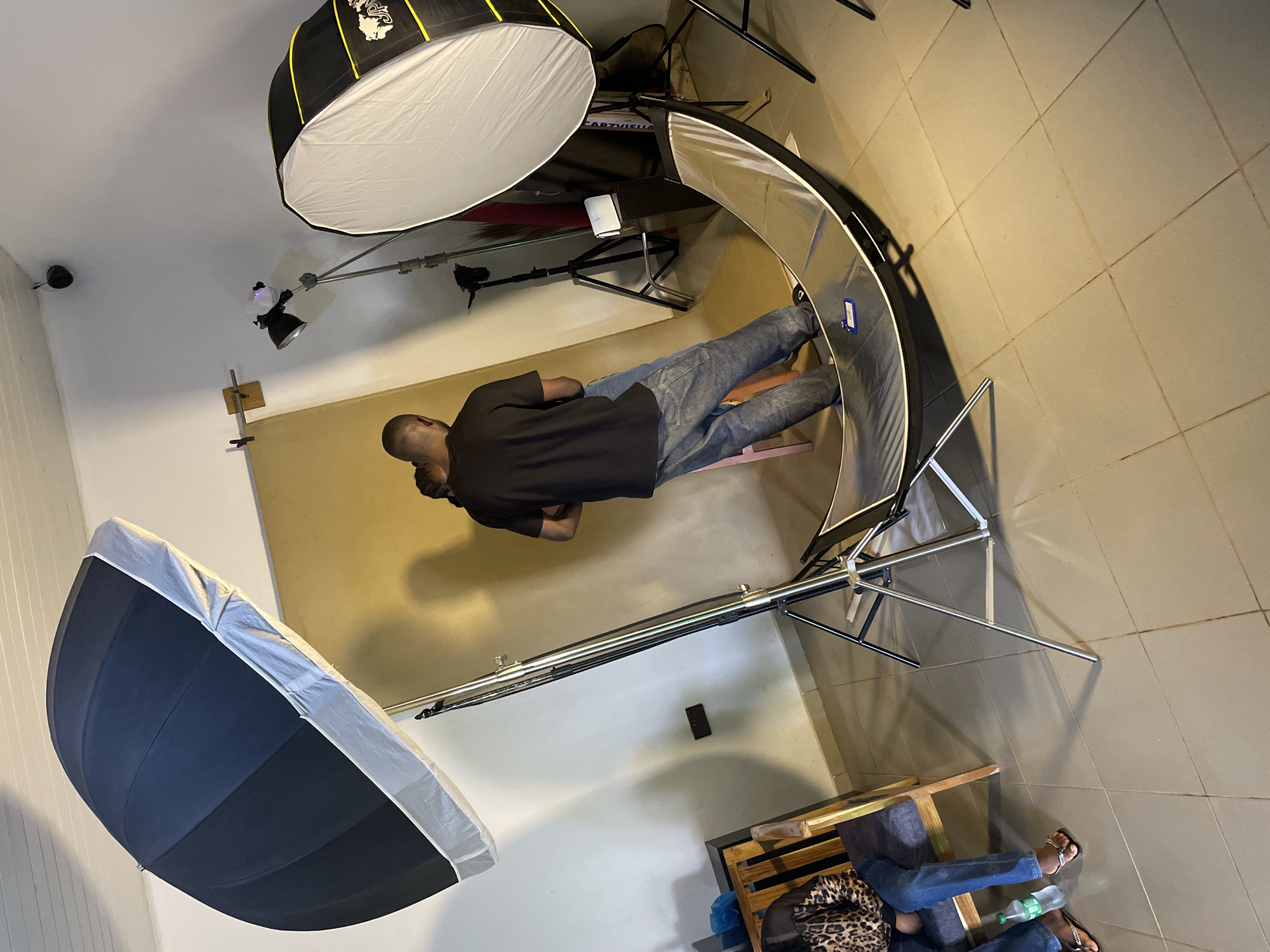This image captures a photography studio setting in Durumi, Nigeria. Two individuals, a man and a woman, are the central subjects, positioned in front of a tan backdrop. The man, wearing a black t-shirt and denim jeans, stands with his back to the camera, appearing to interact with the woman. The woman is partially visible, facing away from the camera.
The studio is equipped with professional lighting gear. A large, dark umbrella-style softbox is positioned to the left, angled downwards. To the right, a large octagonal softbox is set up. A curved reflector lies on the floor in the foreground, reflecting the studio lights. Other equipment visible includes light stands and a tripod.
In the background, a wooden chair with a patterned cushion is occupied by a person wearing jeans and flip-flops, with a plastic bottle of water beside them. The walls of the studio are painted white, and the floor is tiled. The overall impression is a functional space prepared for a photoshoot.