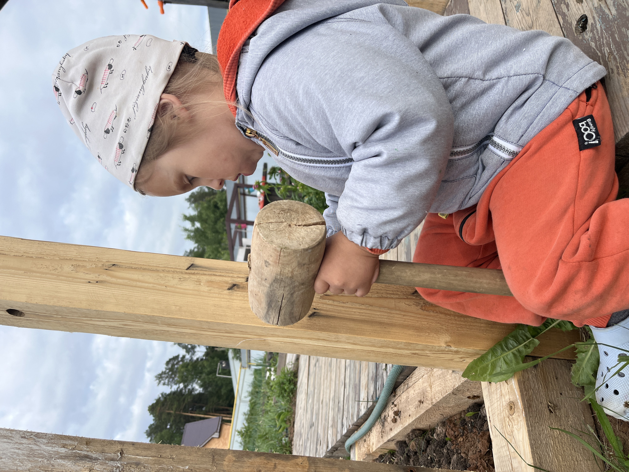 A young child, wearing a grey hooded jacket with an orange lining, orange sweatpants, and a bandana with a dog pattern, is sitting on a wooden surface. The child is holding a wooden mallet with both hands, appearing to be engaged in some sort of construction or play. The background shows a wooden structure, greenery, and a cloudy sky, suggesting an outdoor setting. The location is identified as Chaikovsky, Russia.