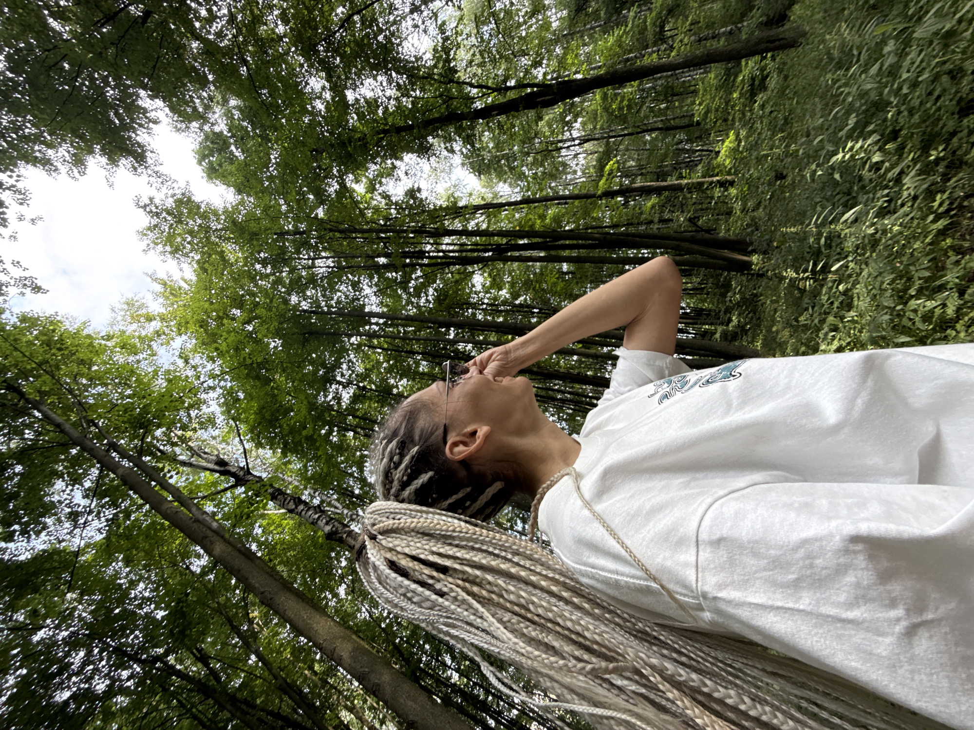 In Rusanovo, Russia, a woman with long, blonde braided hair stands in a lush green forest, looking to her right with a hand near her face. She wears a white t-shirt and glasses. The dense forest surrounds her with tall trees and abundant foliage, suggesting a natural, outdoor setting. The sunlight filters through the leaves, creating a dappled effect on the forest floor. The woman's posture and gaze indicate she is observing something within the forest.
