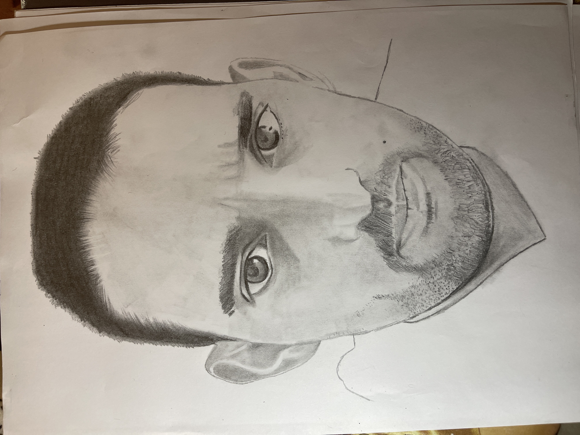 A pencil drawing of a man's face on white paper. The man has a shaved head with short, dark hair, visible eyebrows, and realistic eyes. He also has a beard. There are no other visible details or context about a location in Jos, Nigeria.