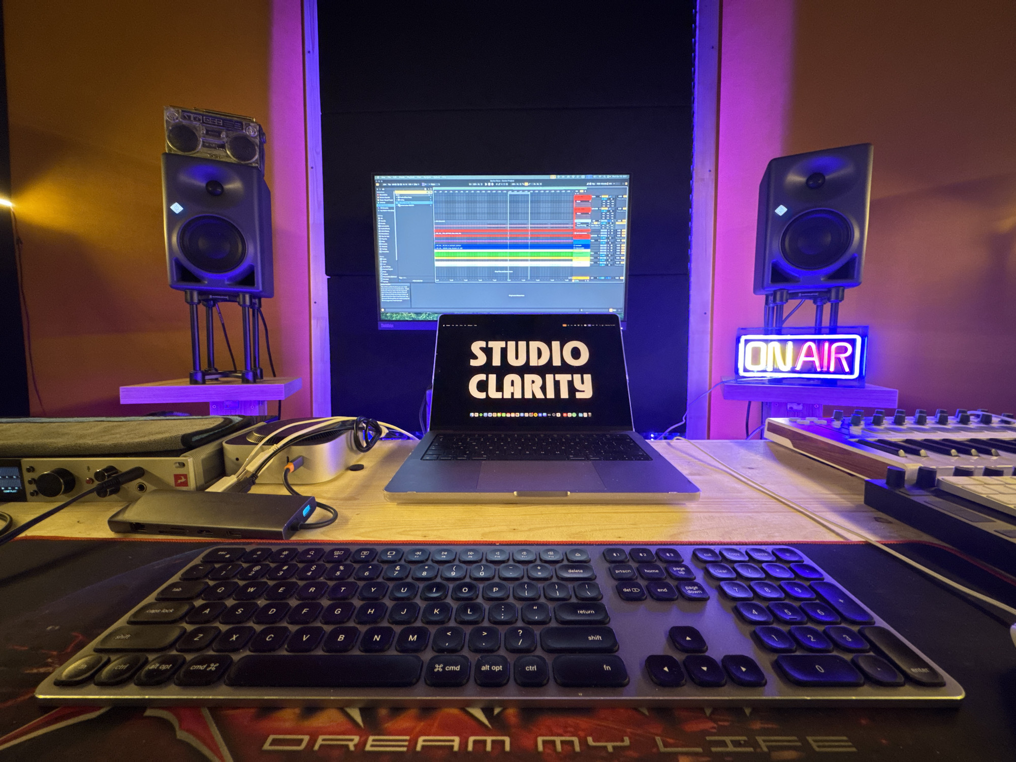 The image shows a music production setup in a studio environment. A keyboard sits in the foreground on a mousepad with the words "DREAM MY LIFE" visible. Behind the keyboard is a desk with a laptop displaying "STUDIO CLARITY" on the screen, positioned in front of a larger monitor displaying music production software. On the desk are various pieces of audio equipment, including an audio interface and a hard drive. To the left and right of the monitor are studio monitor speakers, one with a boombox on top and the other with a neon "ON AIR" sign underneath. The walls of the studio are visible in the background, with the left wall appearing yellow and the right wall a lighter color. Purple ambient lighting is present in the space. The location is not mentioned or discernible from the image.