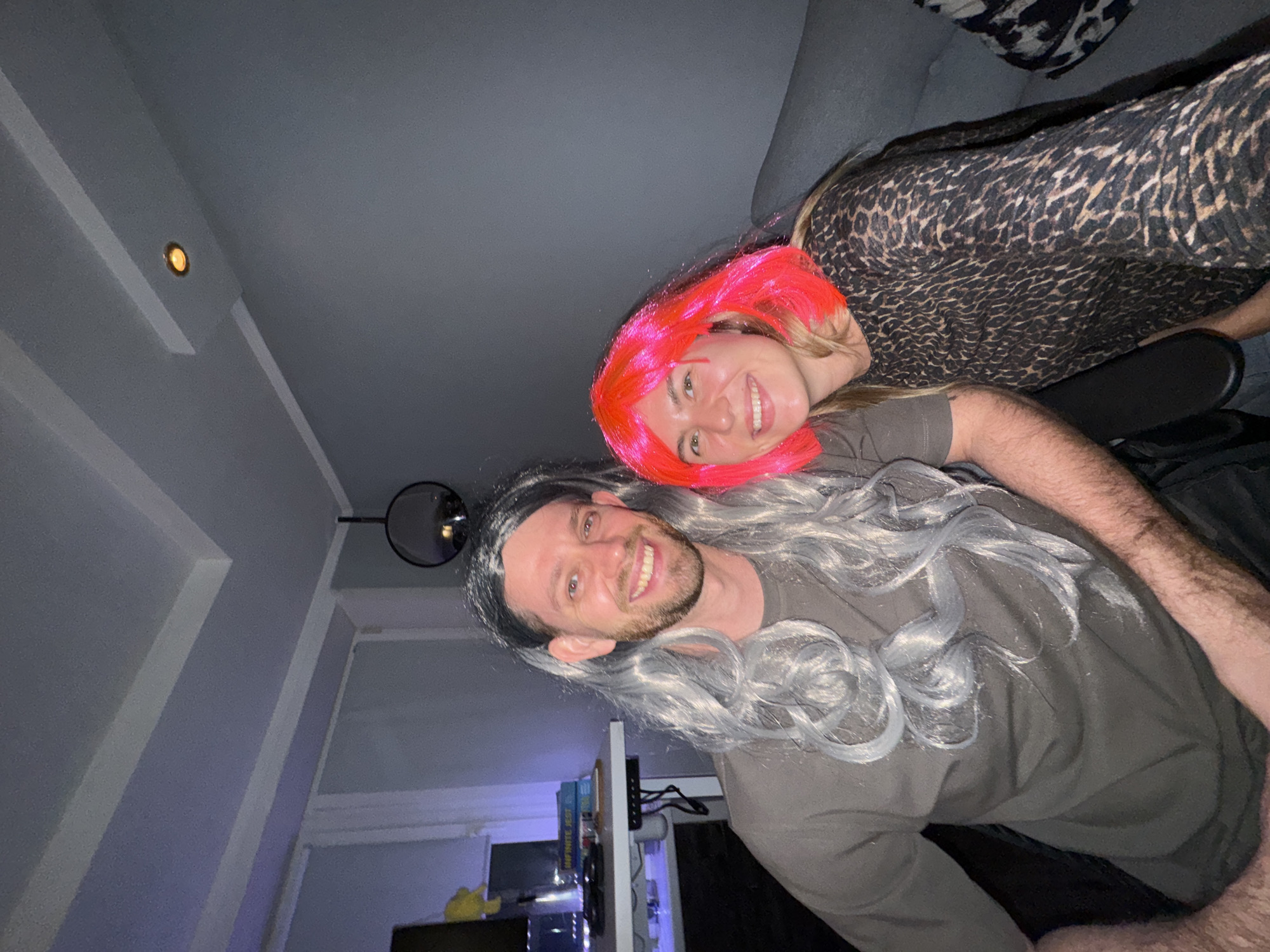 In an indoor setting, possibly a living room or office, two individuals are posing for a photograph. On the left, a man with a beard is wearing a long, grey, curly wig. He is dressed in a dark grey t-shirt. Beside him on the right, a woman is wearing a bright pink wig and a long-sleeved top with a black and brown leopard print pattern. Both individuals are smiling and appear to be looking at the camera. The background reveals a grey wall, a ceiling with recessed lighting and decorative molding, and a glimpse of a desk with books on the left. The overall atmosphere suggests a casual or festive occasion.