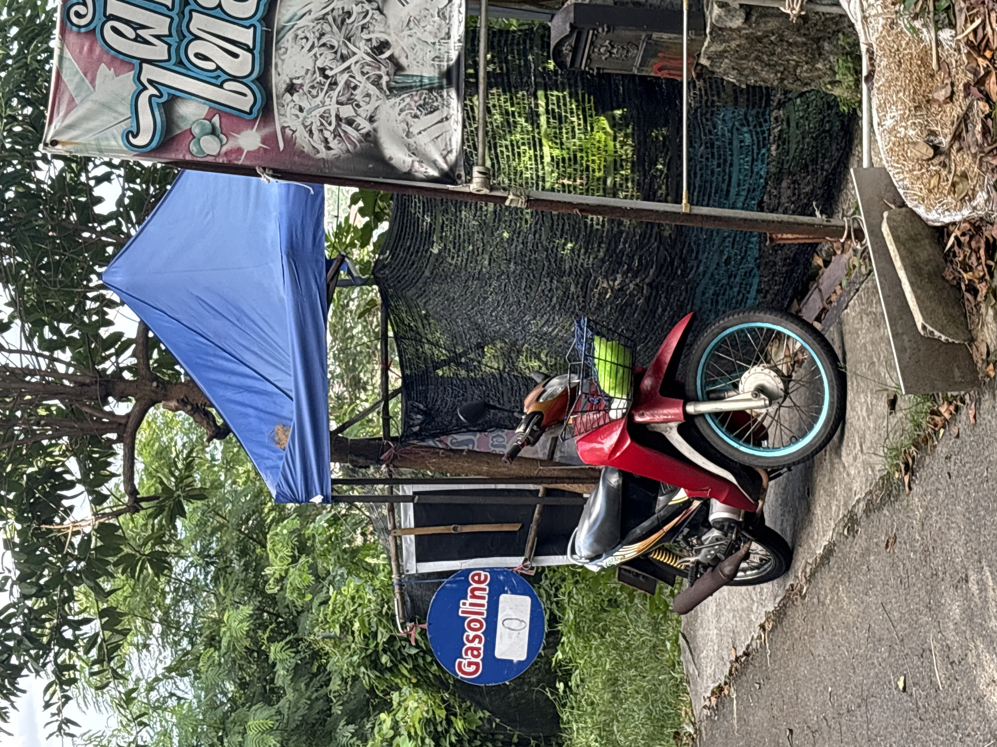 A red motorcycle is parked on a concrete path in front of a blue tent. A round blue sign with the word "Gasoline" in white letters and a white box with the number "0" is hanging to the left of the motorcycle. A vertical banner with Thai writing and an image of noodles is visible to the right of the tent. The background consists of lush green foliage and trees. The scene appears to be a small roadside stall.
