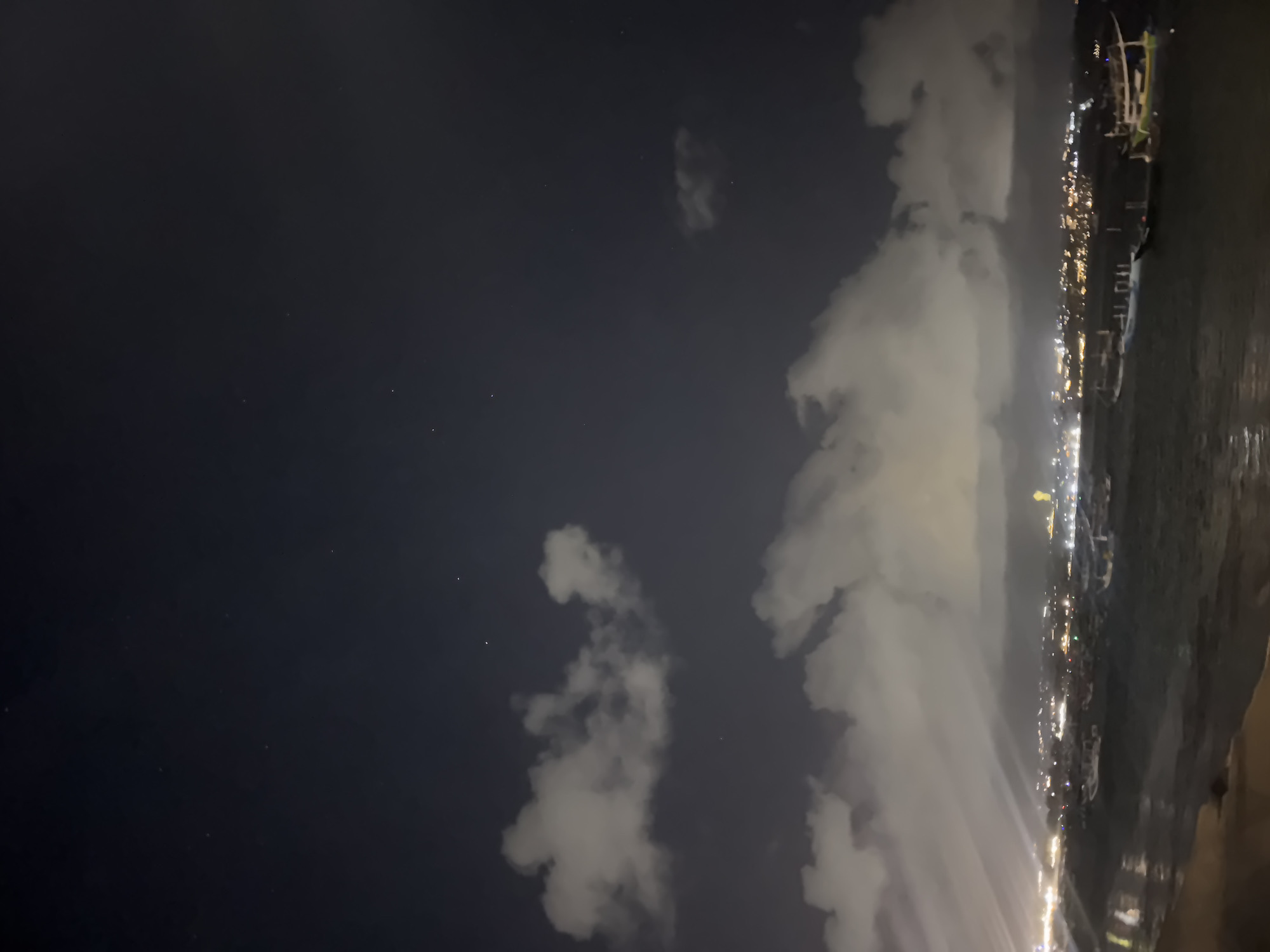 Captured at night in Kuta, Indonesia, the photograph presents a view of the coastline. The night sky is dark with a few scattered clouds. The cityscape is visible along the coast, illuminated with bright lights. The sea lies adjacent to the city, and the edge of the shoreline is visible.
