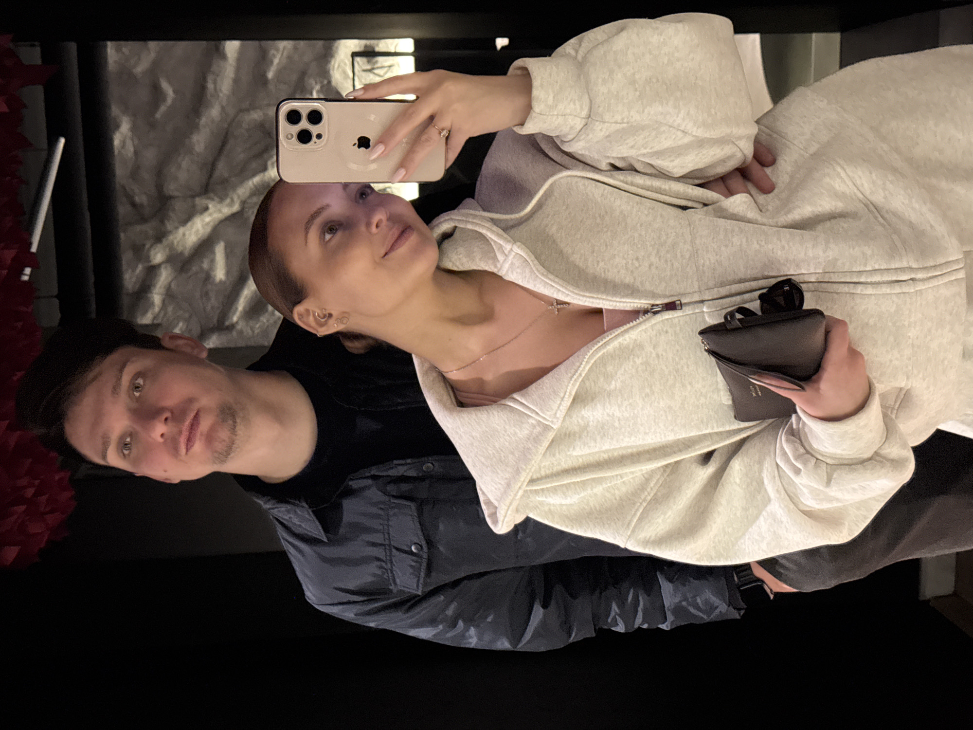 The image shows two individuals taking a selfie. A woman is holding a smartphone, likely an iPhone, to capture the picture. She is wearing a light grey hoodie and a delicate necklace. A man stands beside her, wearing a black shirt and a dark jacket with a visible pocket. The background appears to be a wall with some textures, possibly suggesting an indoor setting. The location and city are not discernible from the image.