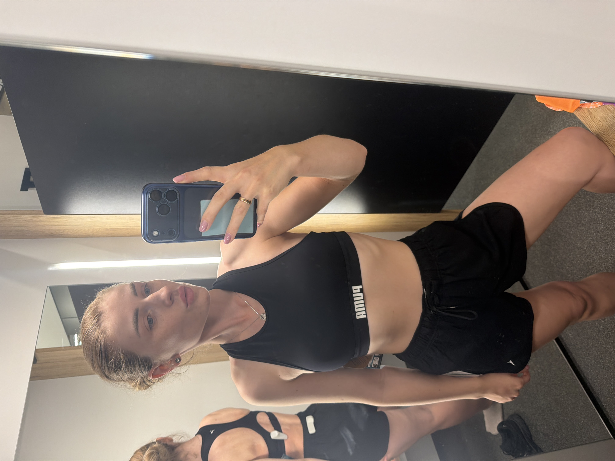 A young woman is taking a mirror selfie in what appears to be a fitting room. She has blonde hair pulled back in a bun and is wearing a black sports bra with a white Puma logo and black athletic shorts. She is looking at her phone, which is a dark blue iPhone with a camera bump. In the background, a reflection of her back is visible in the mirror, showing her wearing the same outfit. There is a black panel on the wall behind her, and a white vertical light strip to the right. The floor is a speckled gray.