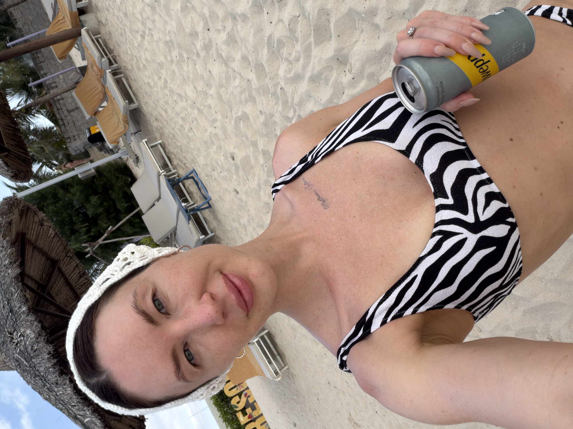 In a sun-drenched beach setting, likely in Dong Ninh Hoa Ward, Vietnam, a woman poses for a selfie. She wears a black and white zebra-print bikini top and a white crocheted headscarf. Her left arm rests on her hip, holding a silver can of Schweppes soda. Her right hand, adorned with a ring, is near the can, displaying long, light-colored nails. A faint tattoo is visible on her upper chest. The background reveals a sandy beach with thatched umbrellas, lounge chairs, and palm trees. The word "RESORT" is partially visible on a yellow sign behind her. The sky is a pale blue with scattered clouds.