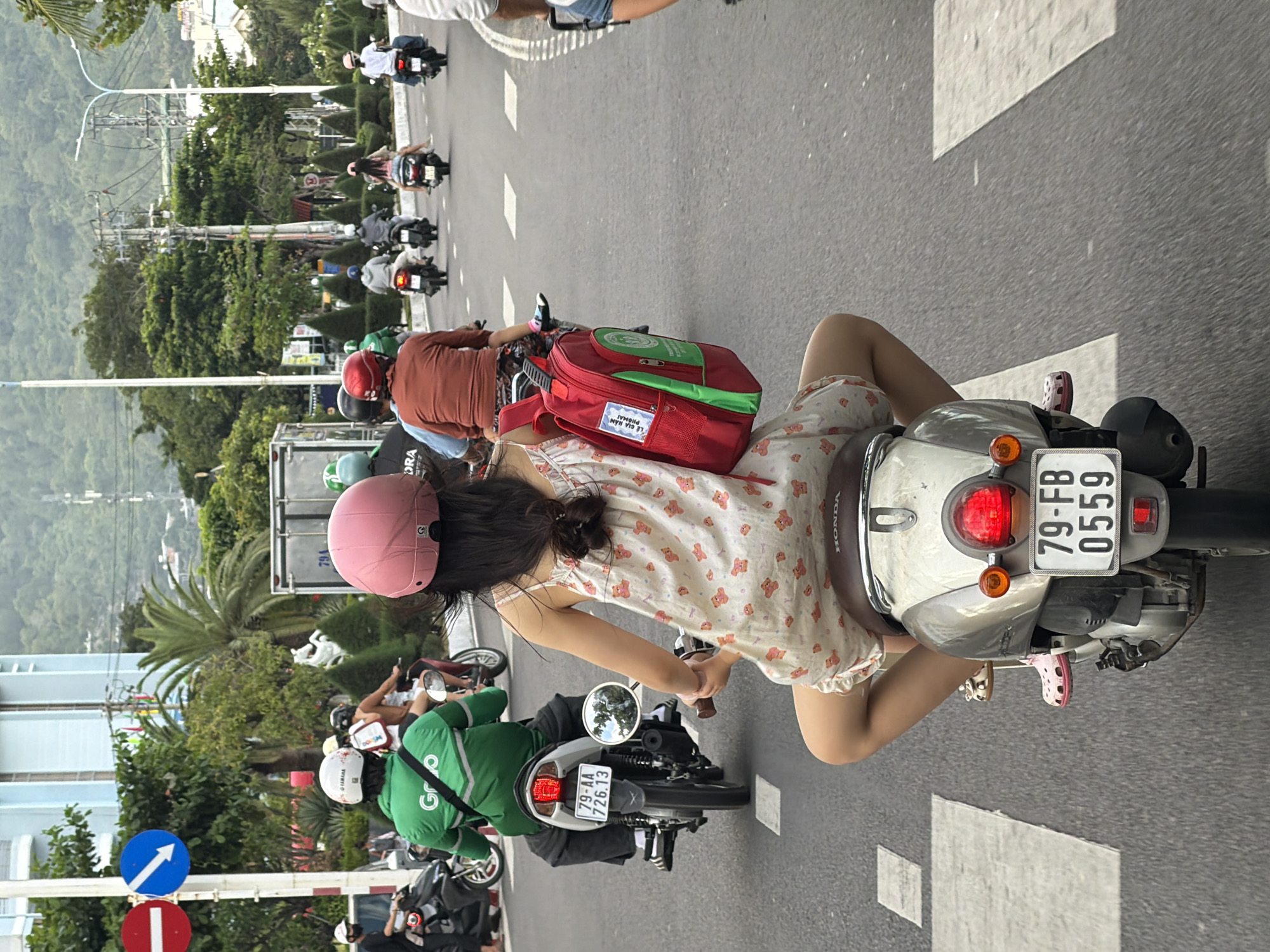A group of motorcyclists are proceeding forward on a city street in Nha Trang, Vietnam. The street is paved and marked with white directional lines. In the background, there are buildings, trees, and power lines. On the left side of the image, a red and blue traffic sign is visible. The primary subject is a young girl wearing a pink helmet and a light-colored dress with a teddy bear pattern. She is riding on the back of a scooter, carrying a red backpack with green accents. She is looking straight ahead. Several other people on motorcycles are also moving forward, with some wearing helmets and others without. One person in front of the girl is wearing a green jacket and a white helmet. Another person further ahead is wearing a brown shirt and a red helmet. The weather appears overcast.
